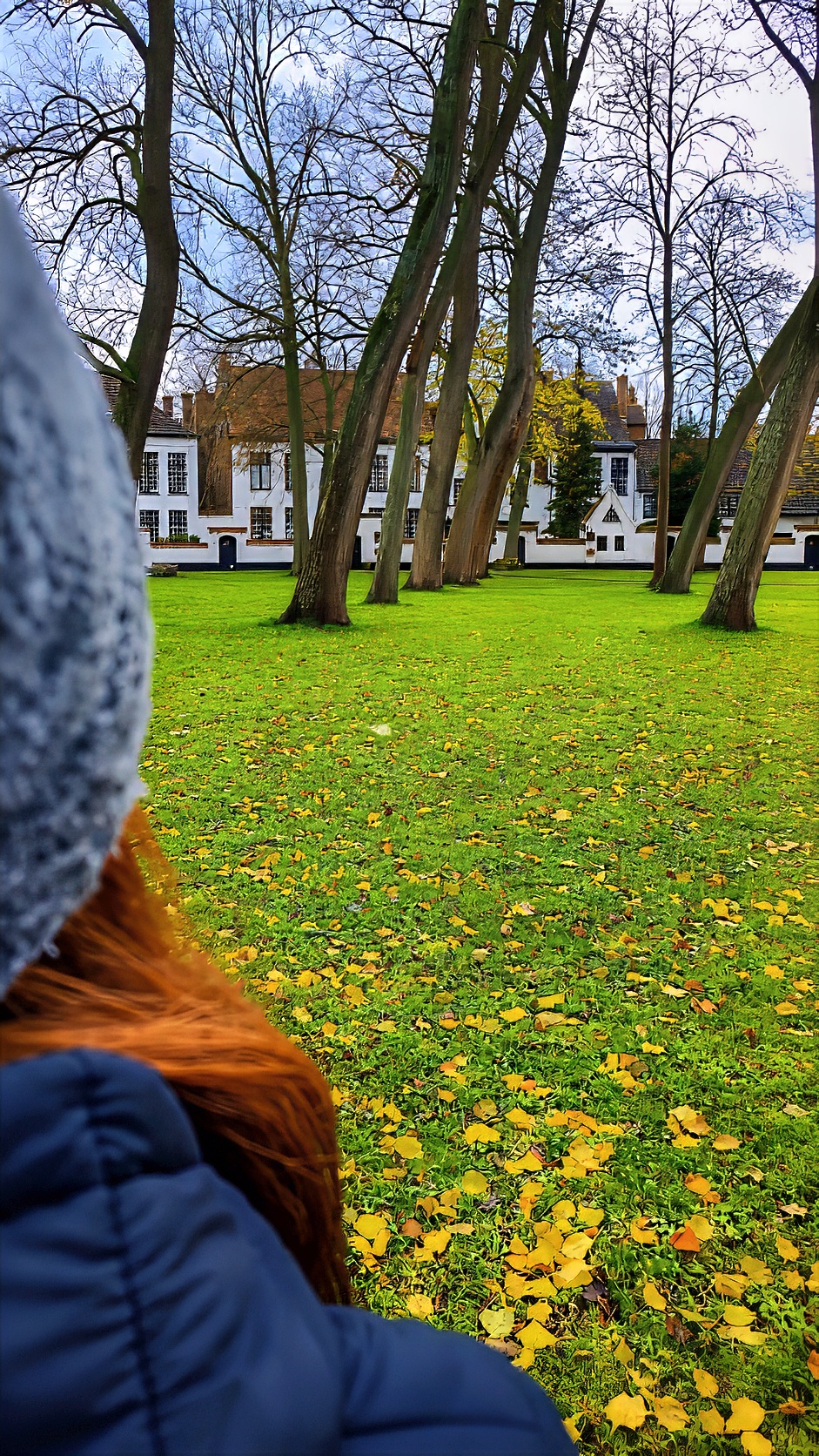 Barbara in a knitted hat, seen from behind, looking across the Beguinage lawn.
