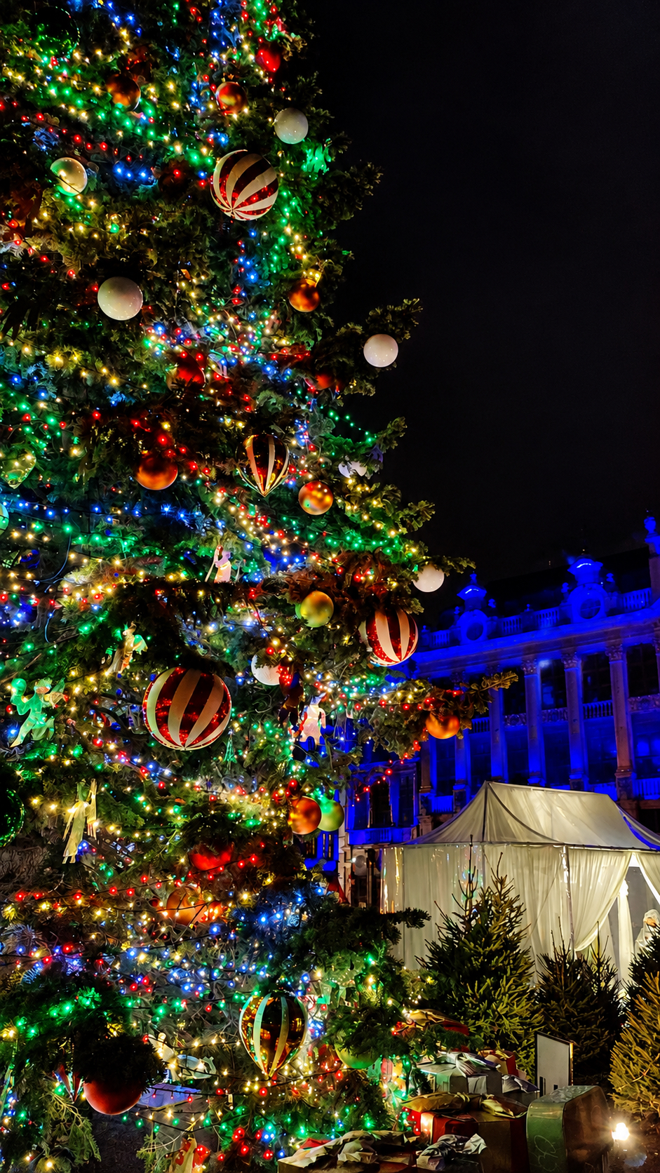 A Christmas tree hung with red baubles, with the lit façades of the Grand-Place behind.