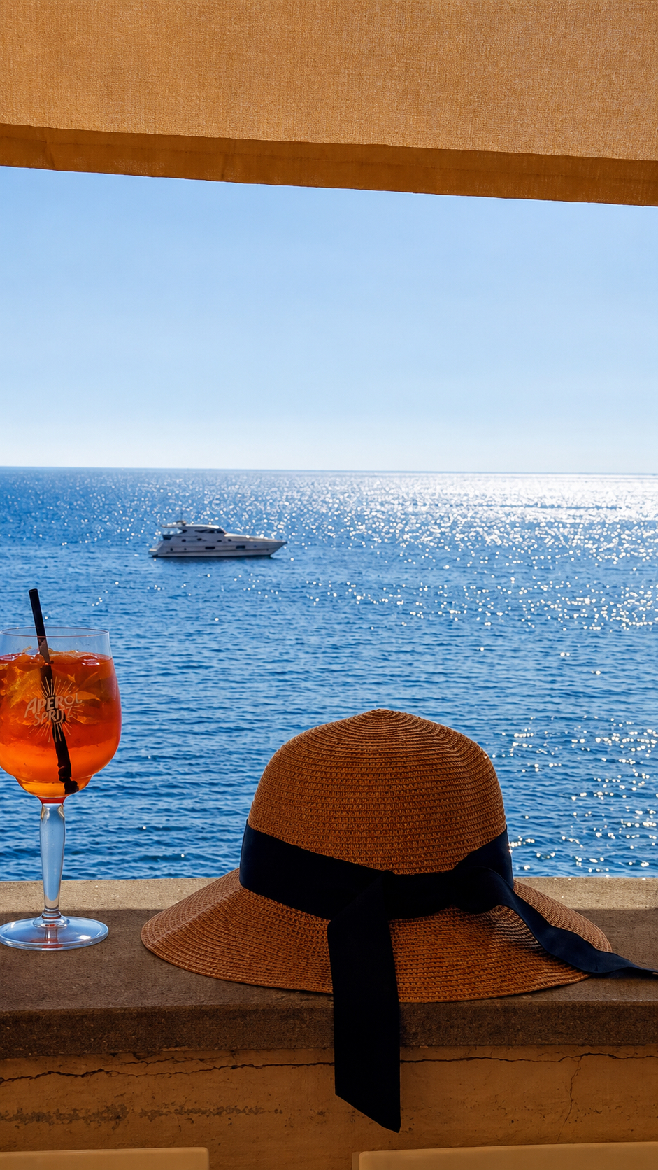 An orange spritz and a straw hat on a sunlit ledge overlooking the sea.
