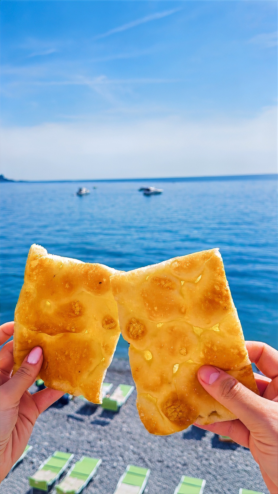 A piece of focaccia held up against the sea, above a row of beach loungers.