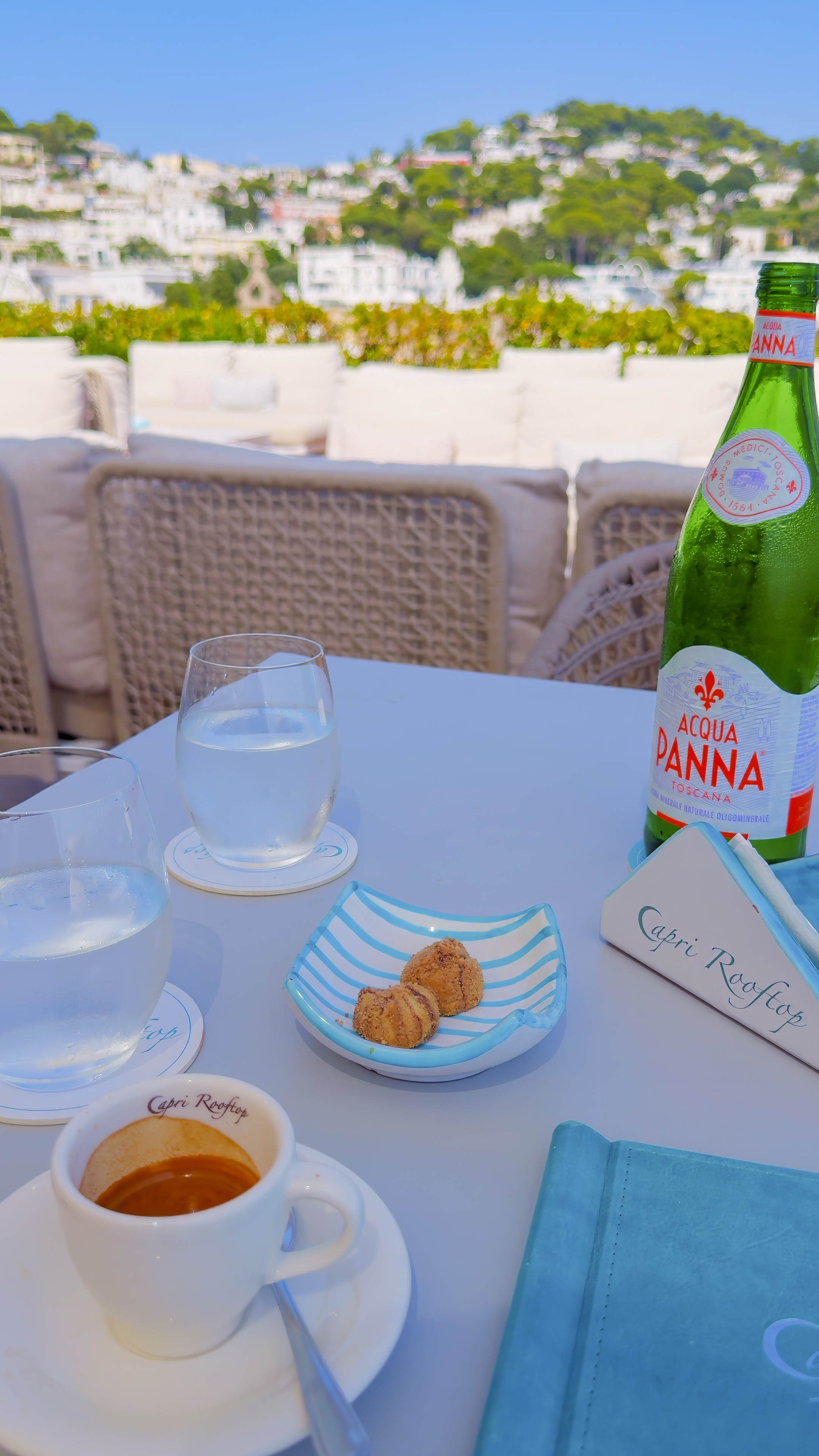 An espresso and a bottle of water on a terrace table, with white houses and the sea beyond.