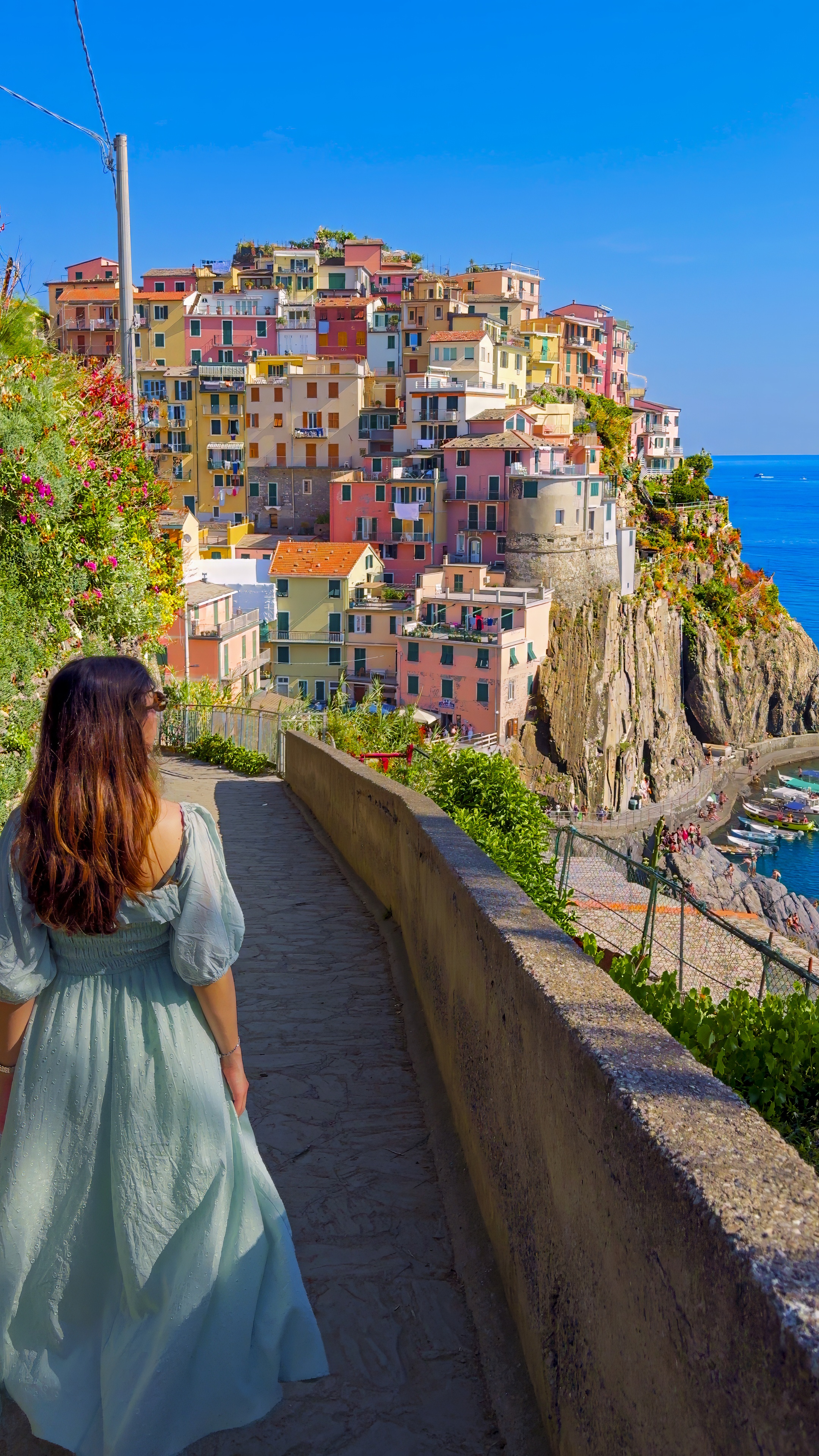 Barbara walking along a stone-walled path, with Manarola stacked on its headland behind.