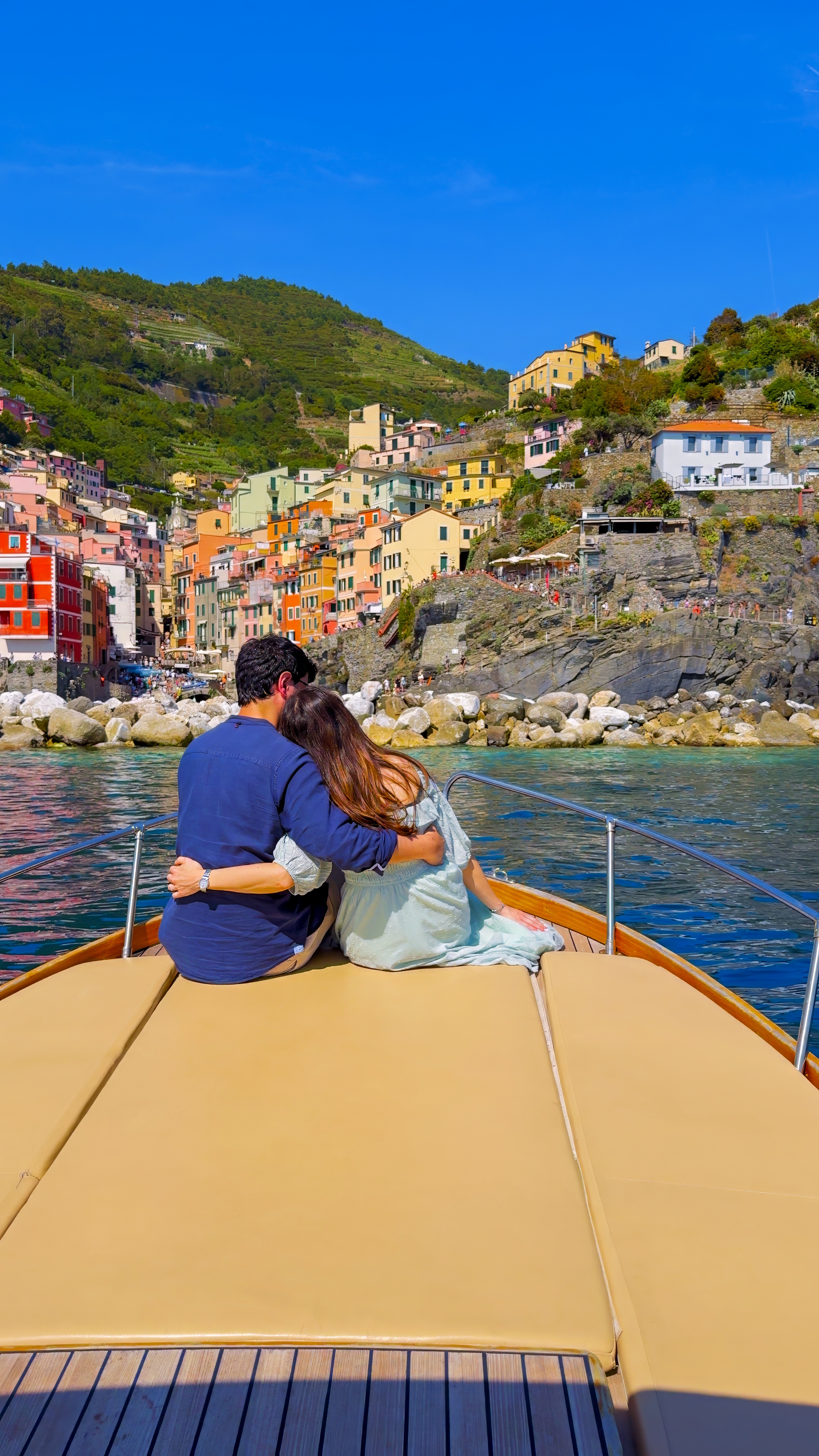 Davide and Barbara sitting on the bow of a boat, facing a village on the cliffs.