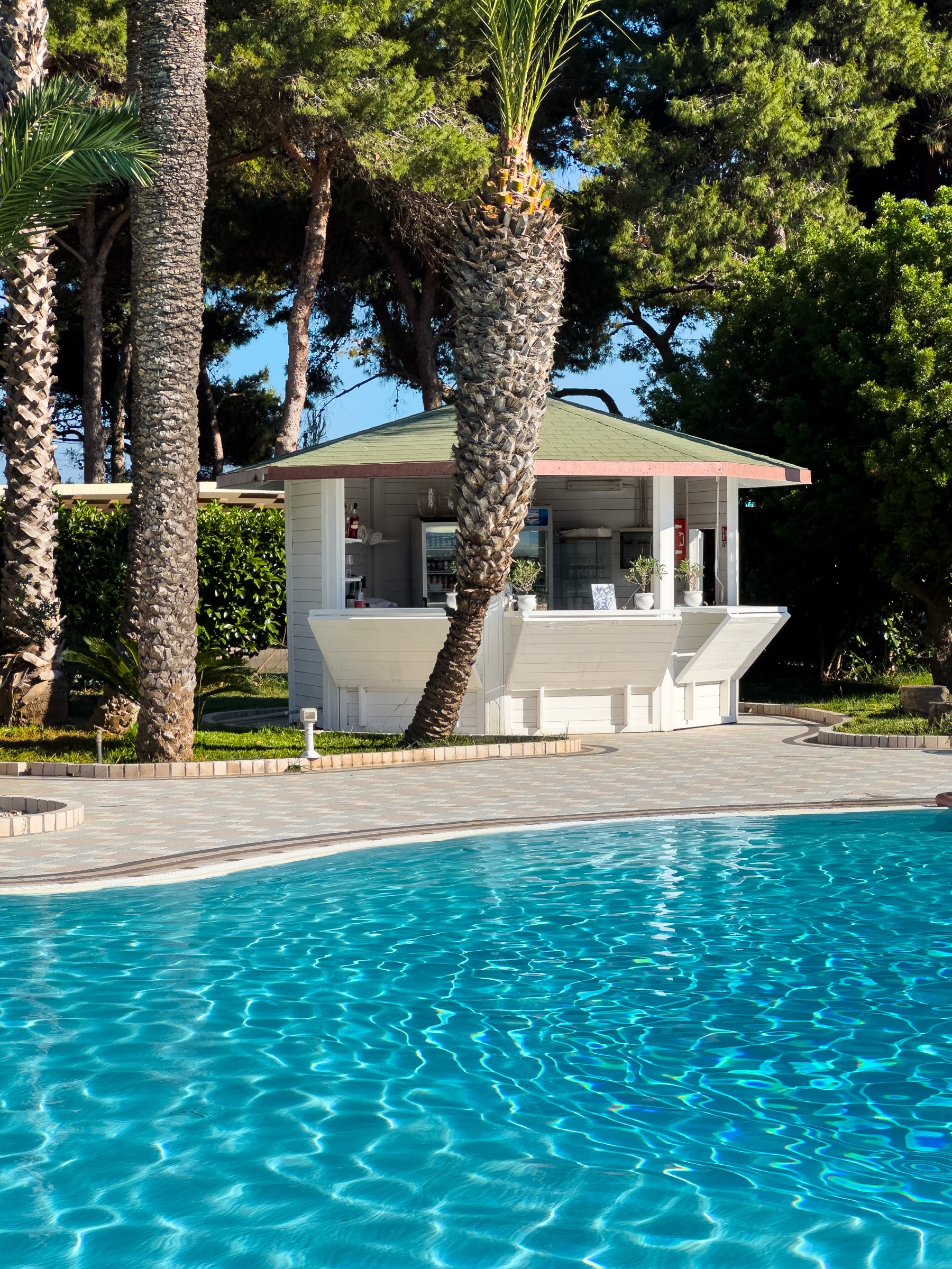 A white pavilion beside the pool, framed by palm trunks and Mediterranean trees