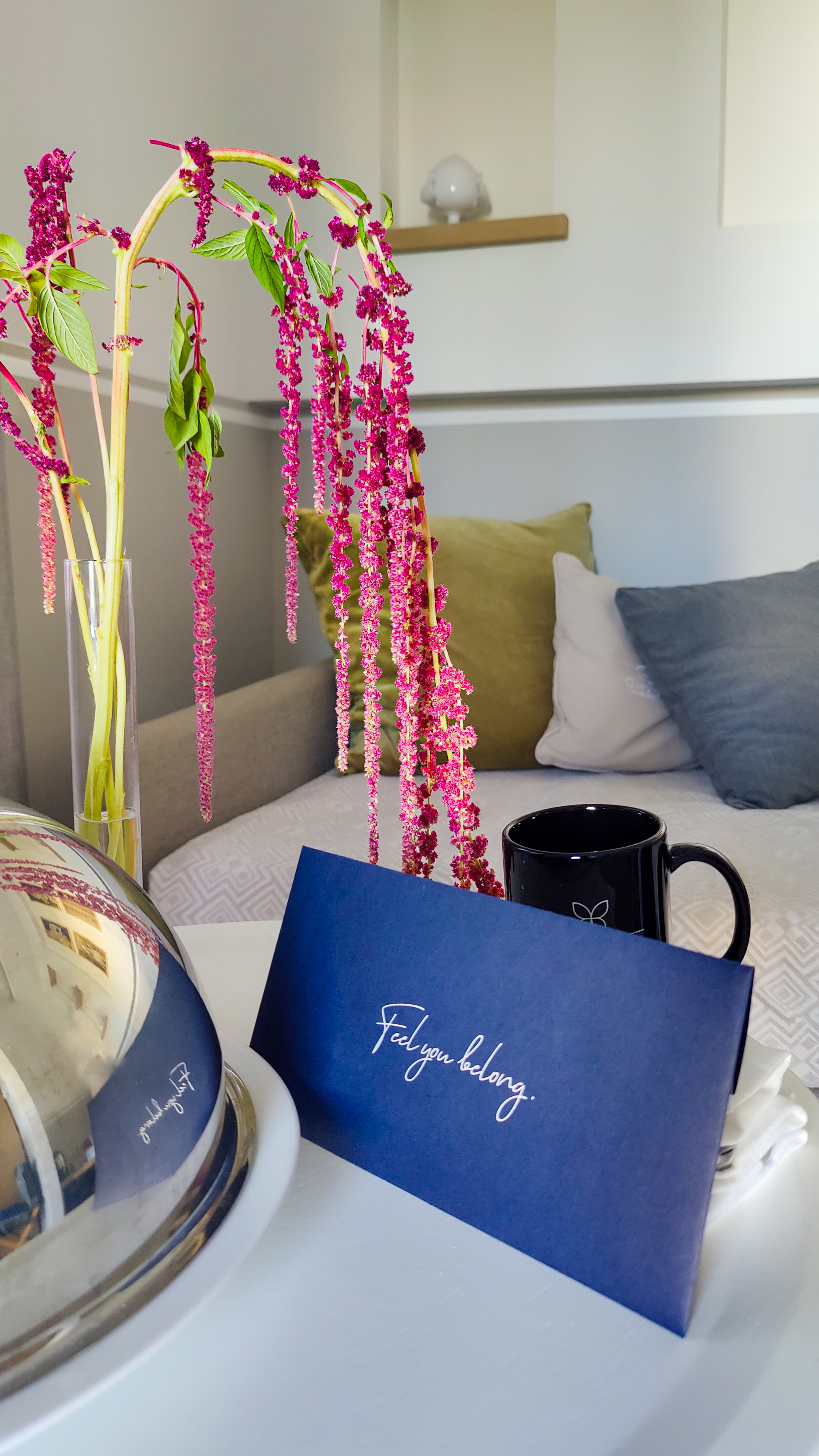 A welcome in the room: a stem of red amaranth, a handwritten card, a dark cup and a covered silver dish