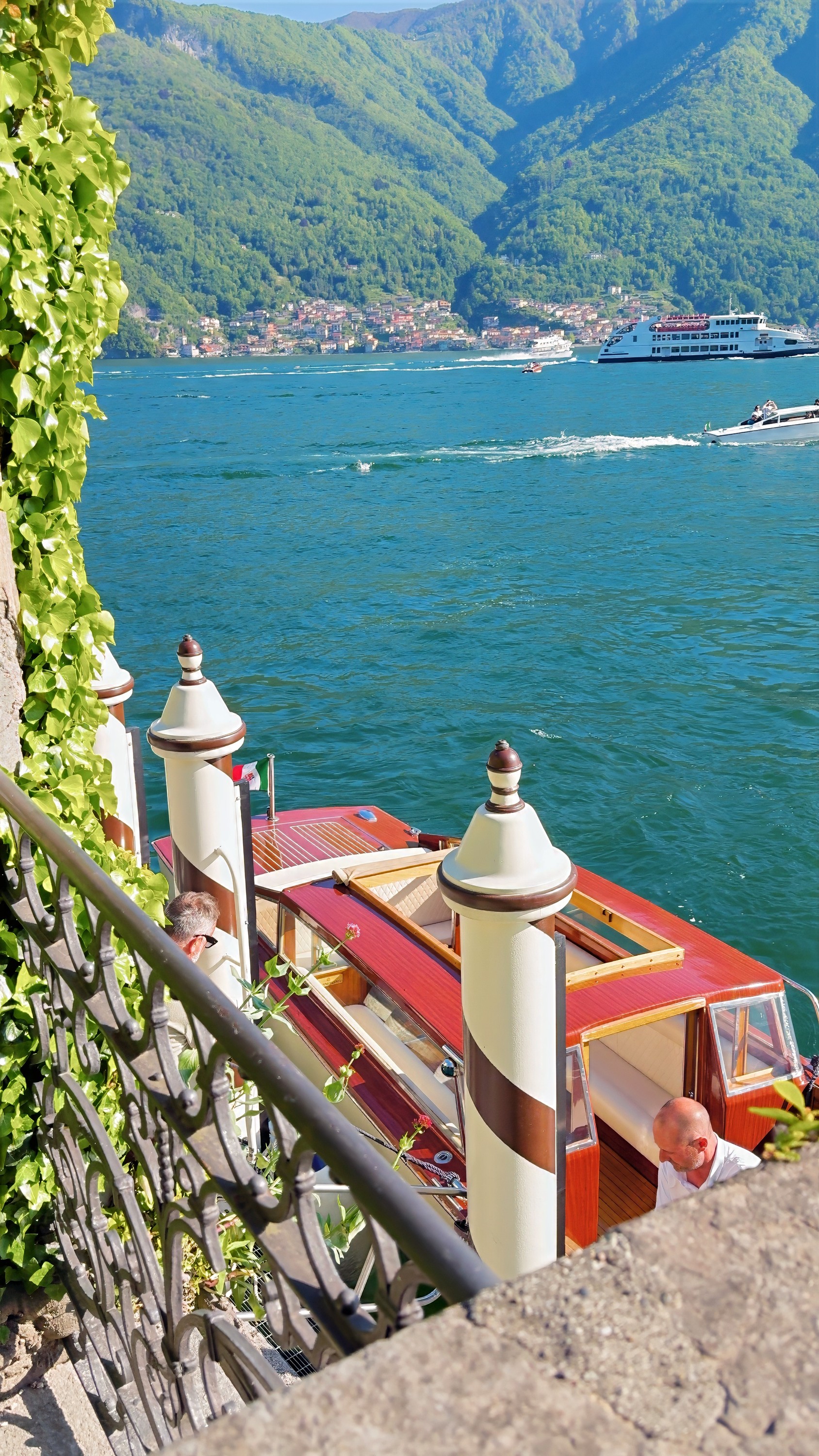 A wooden motorboat moored between striped mooring posts below an iron railing, with a lake ferry and a village across the water.