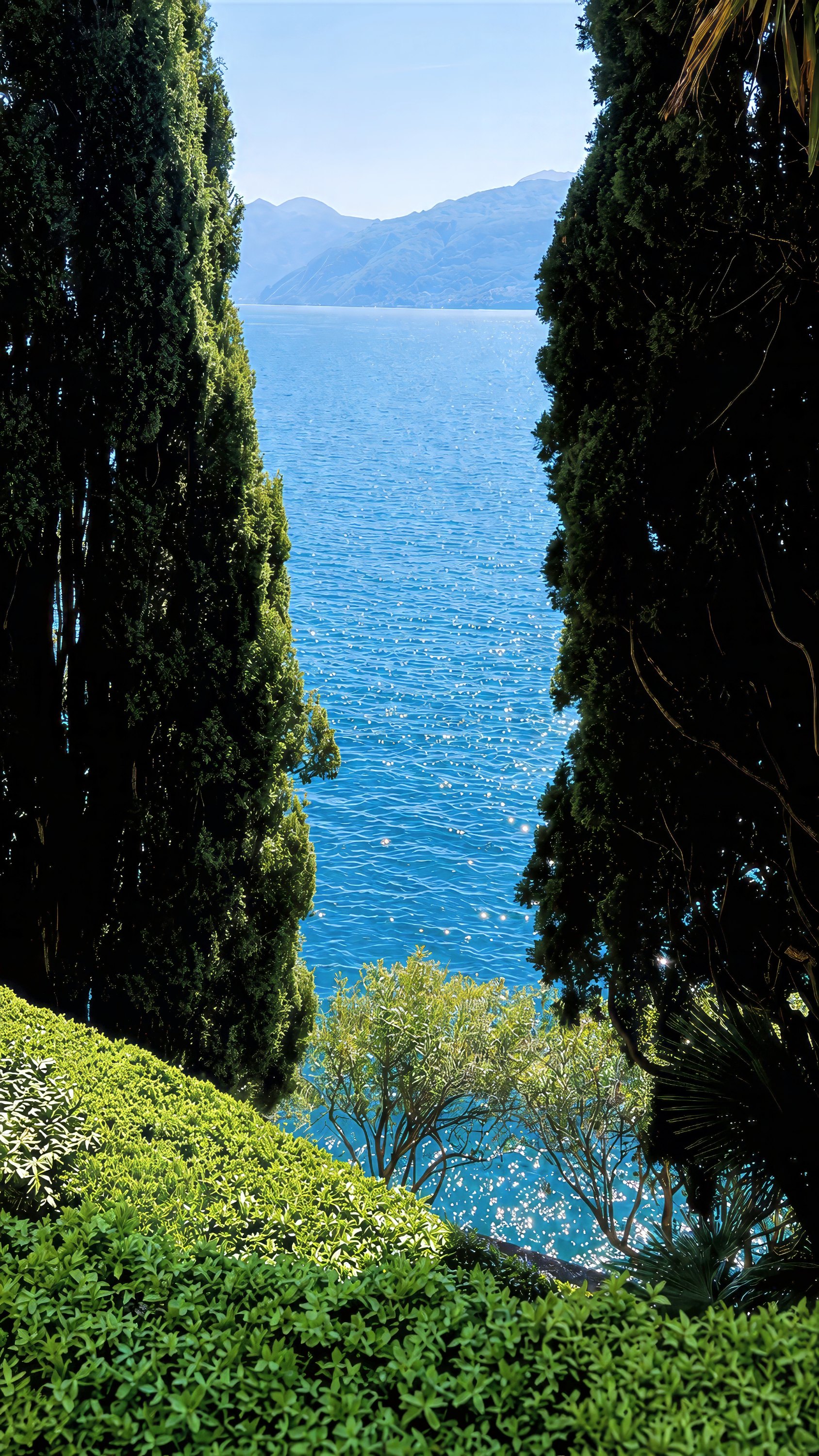 Tall cypresses framing a view of the lake and the mountains on the far shore.