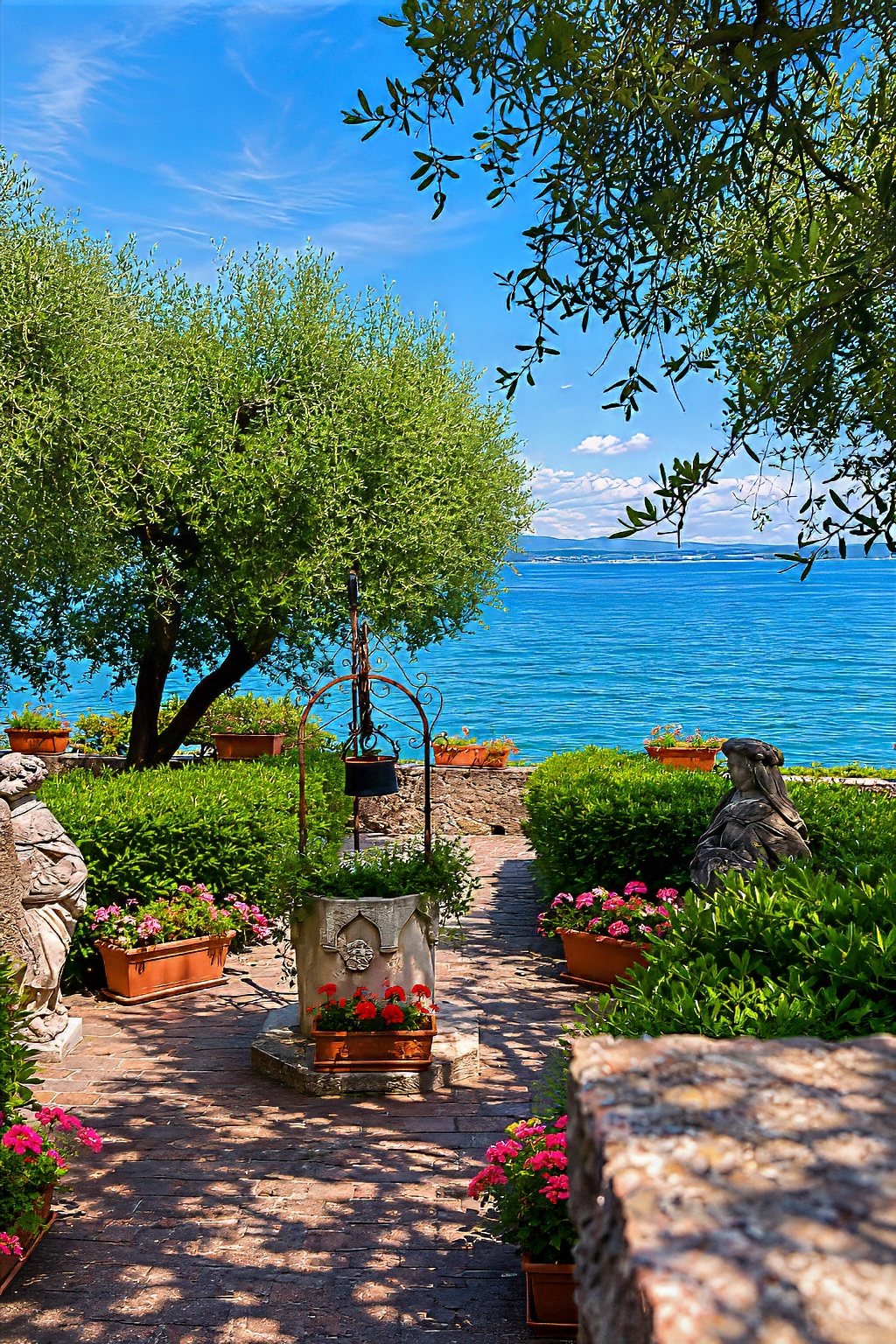 A garden of olive trees, potted flowers and a small stone well above the blue lake.