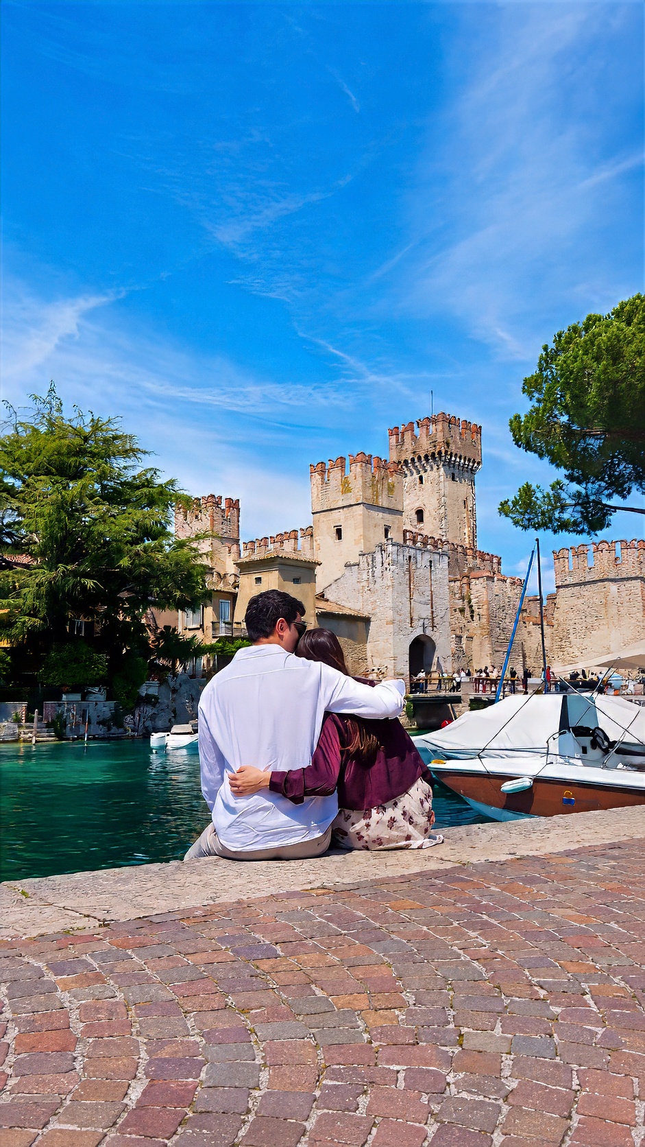 Davide and Barbara sitting on a stone quay, with the towers of a castle rising behind them.