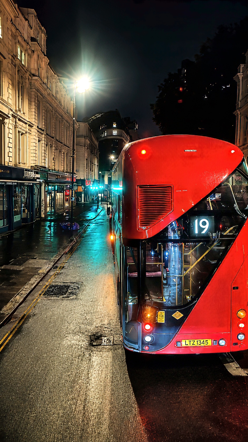 A red double-decker bus on a wet street at night, beside lit stucco buildings.