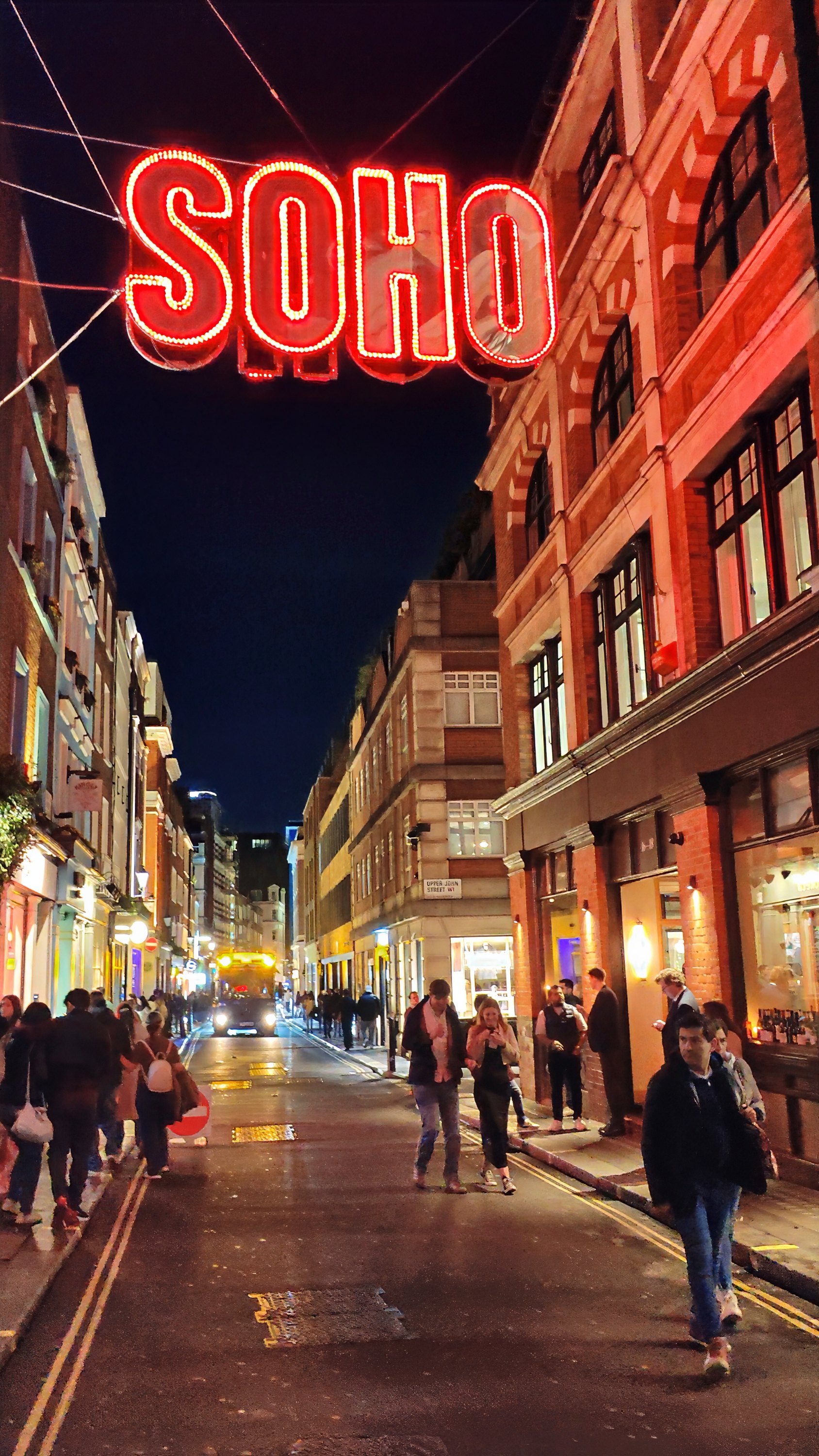 A red neon SOHO sign strung above a street at night, with people walking below.
