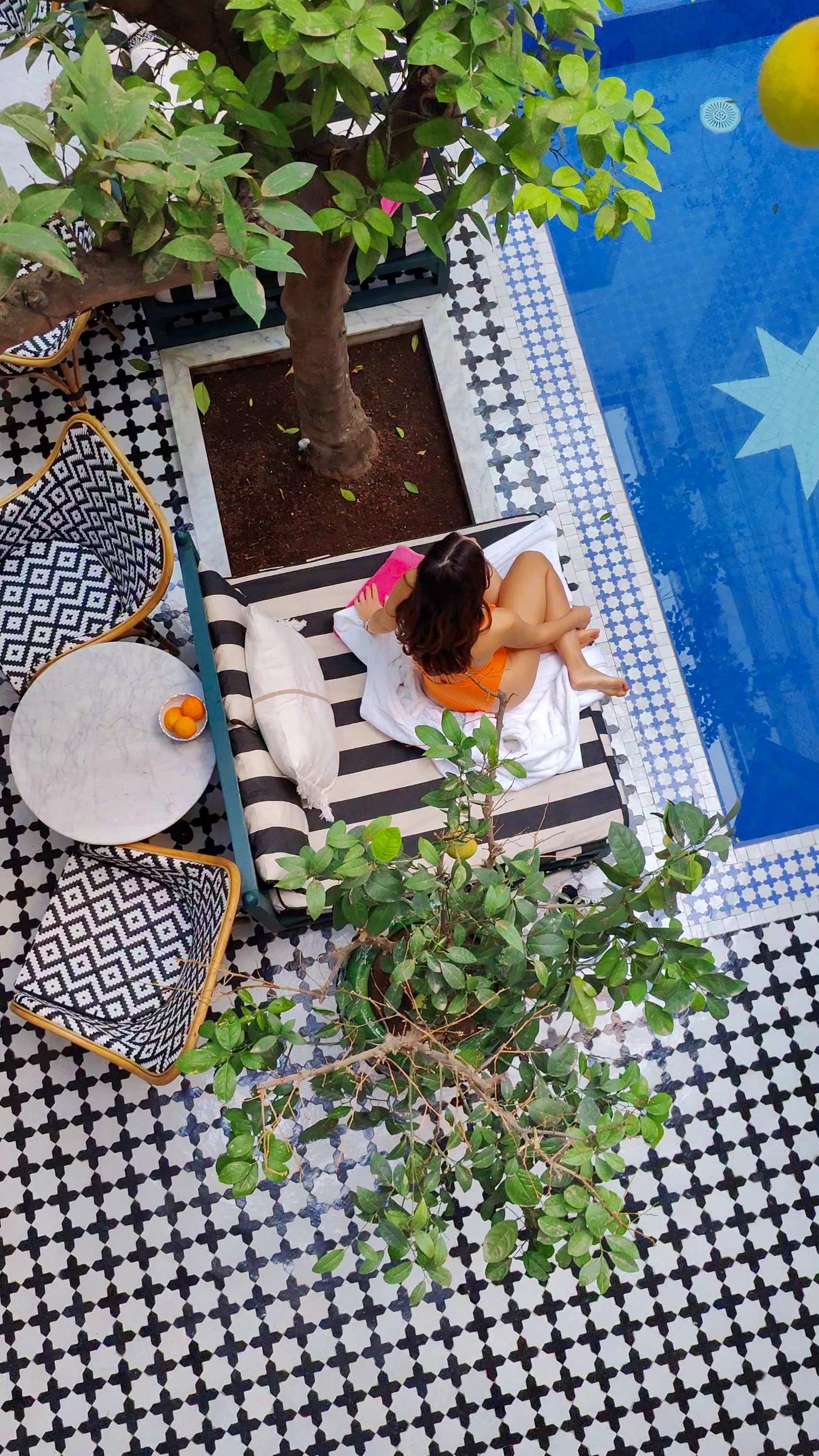 A tiled courtyard seen from above, with a pool, a small tree, cushioned seating and a woman lying on a daybed.