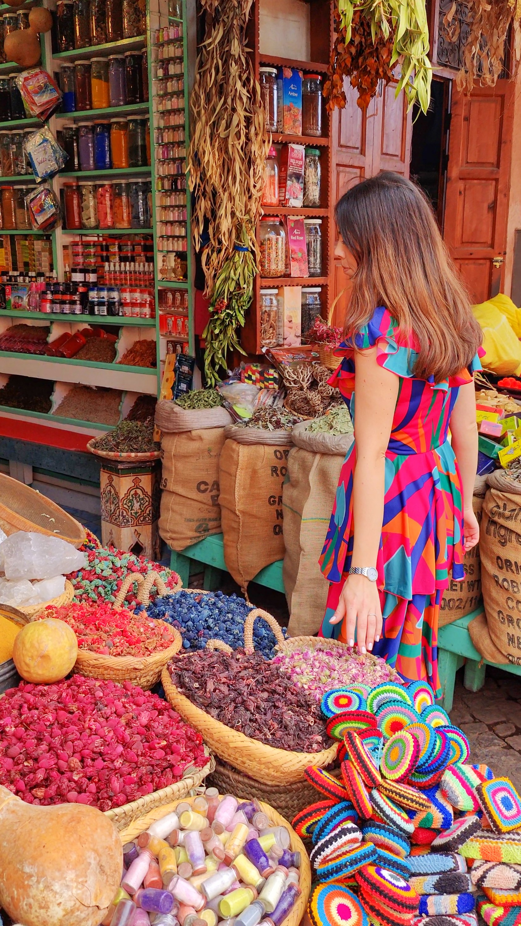 Barbara in a colourful dress beside a stall piled with spices, dried flowers and baskets.