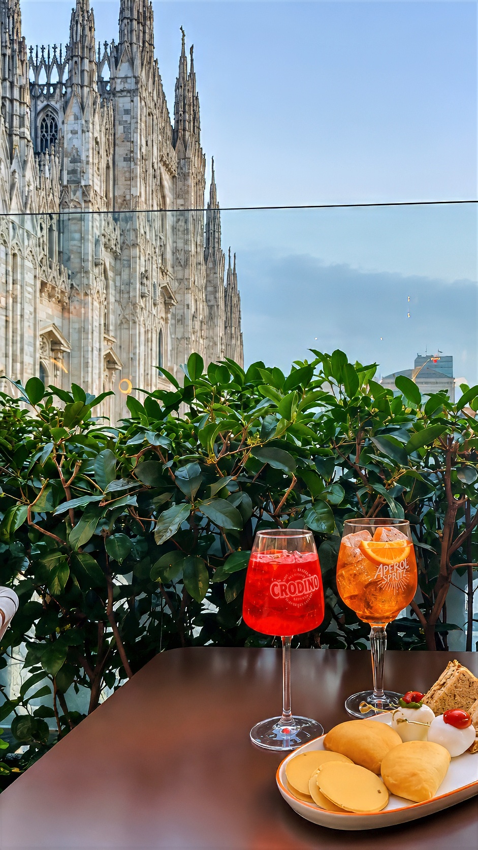 Two spritz glasses and a plate of snacks on a terrace table, with the Duomo spires behind.