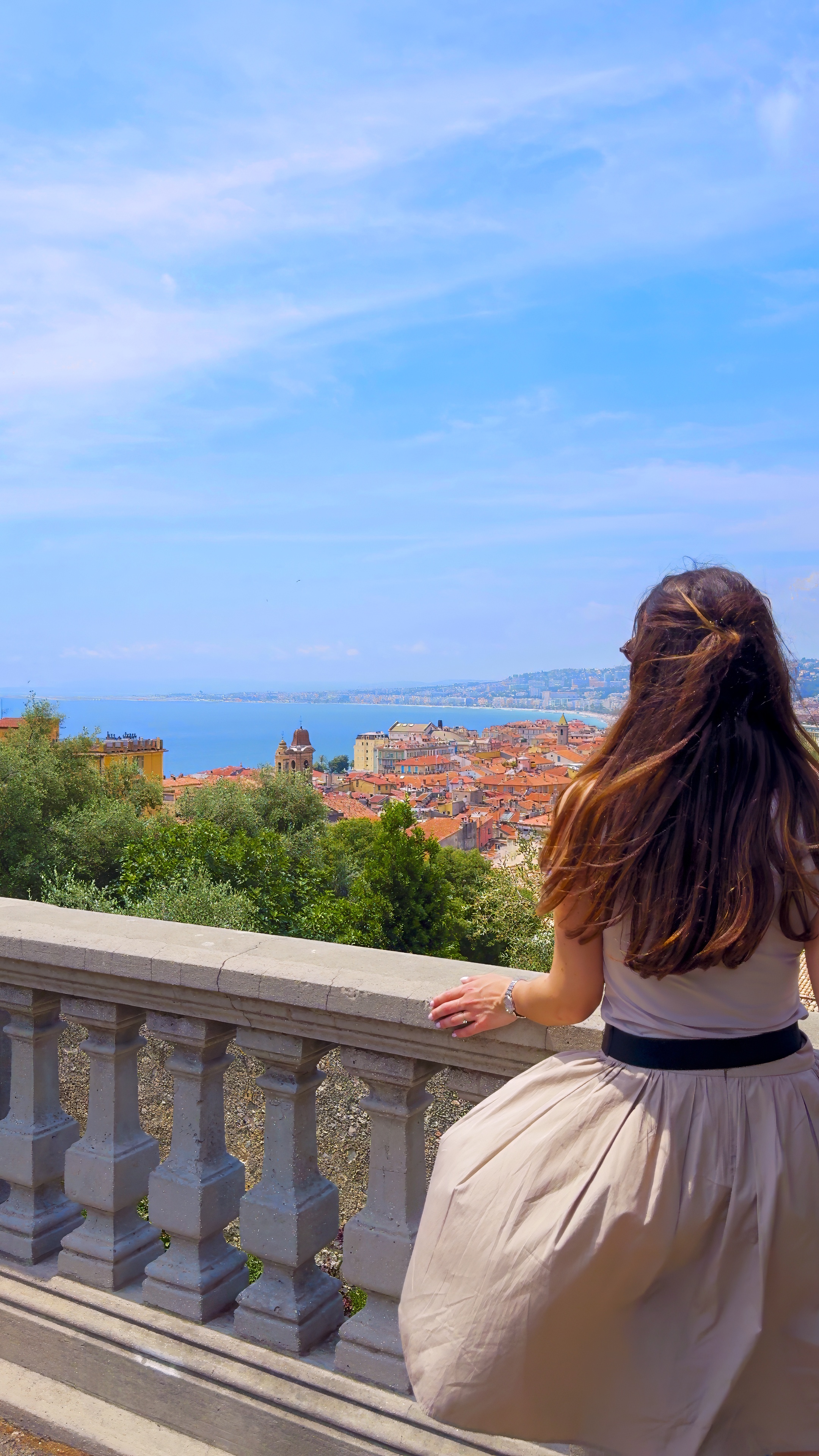 Barbara leaning on a stone balustrade above the city, with the sea and the coast behind.
