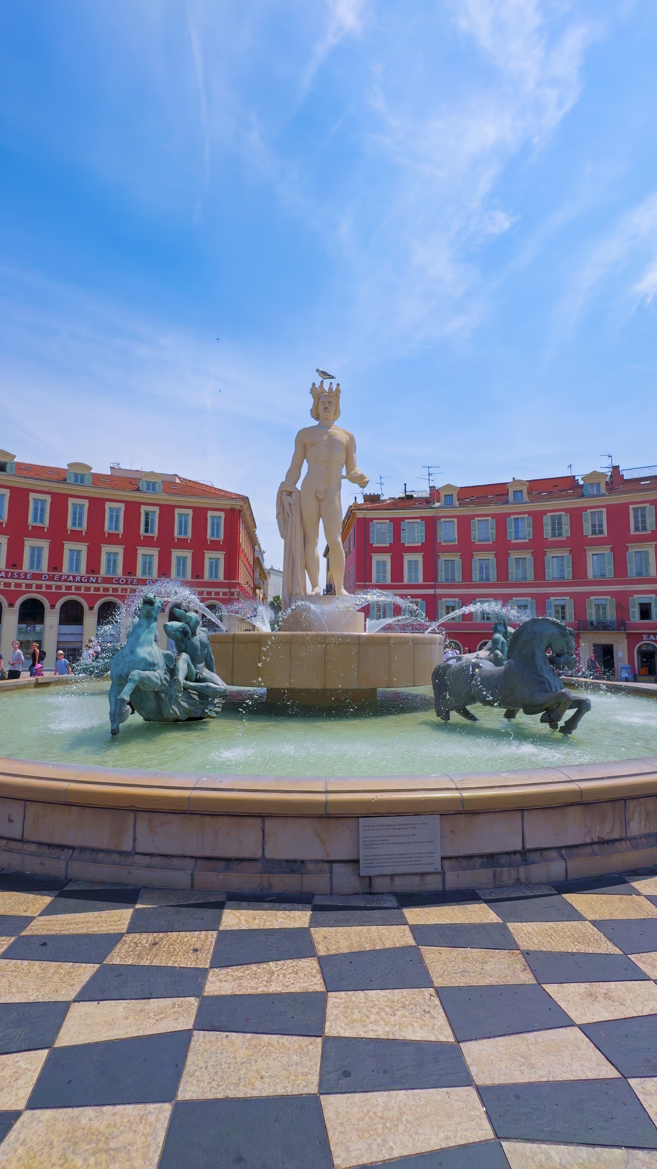 A fountain with a white statue and bronze horses, above black-and-white checkerboard paving, with red façades behind.