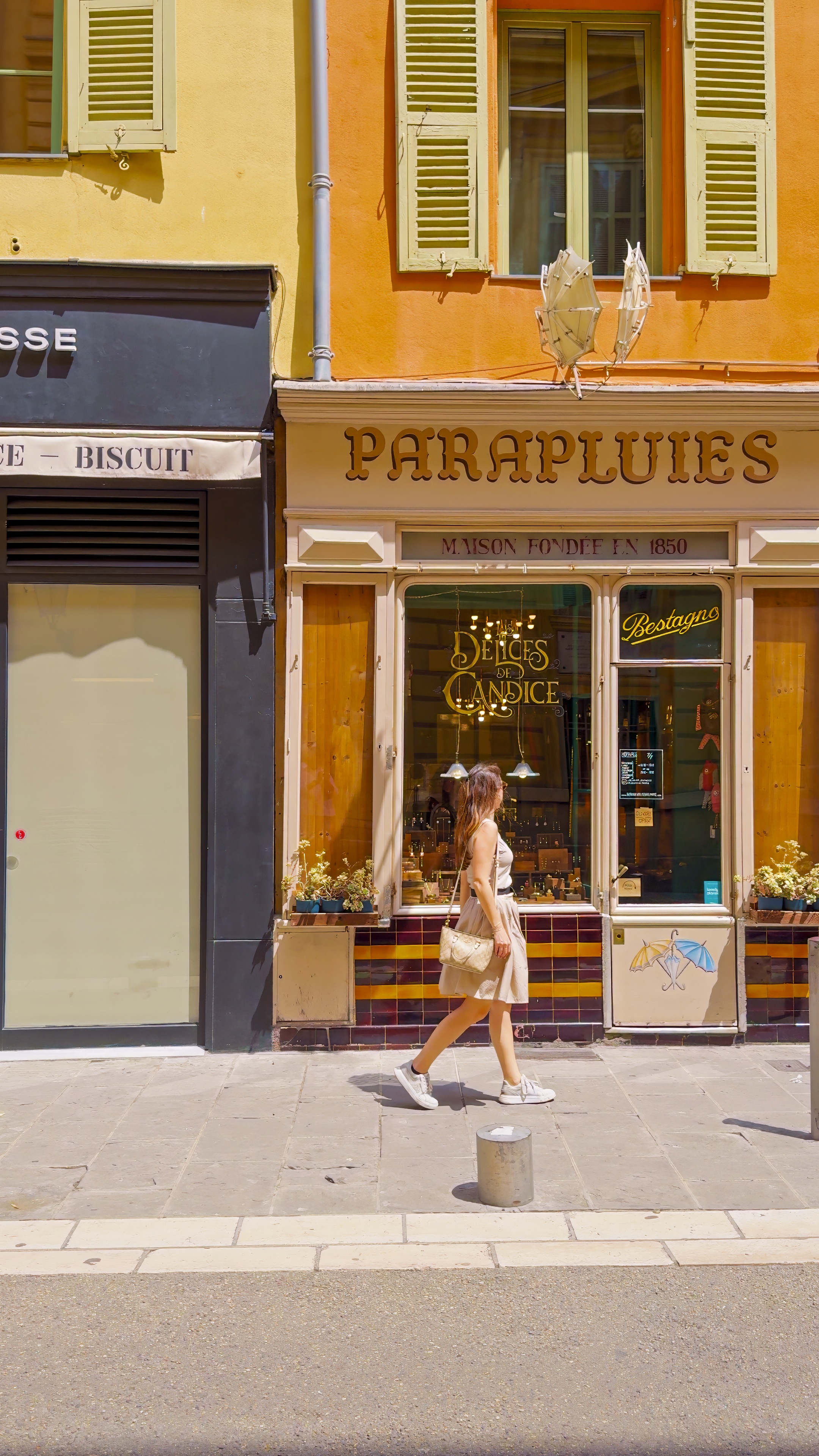 Barbara walking past an ochre shopfront with the word Parapluies above the window.