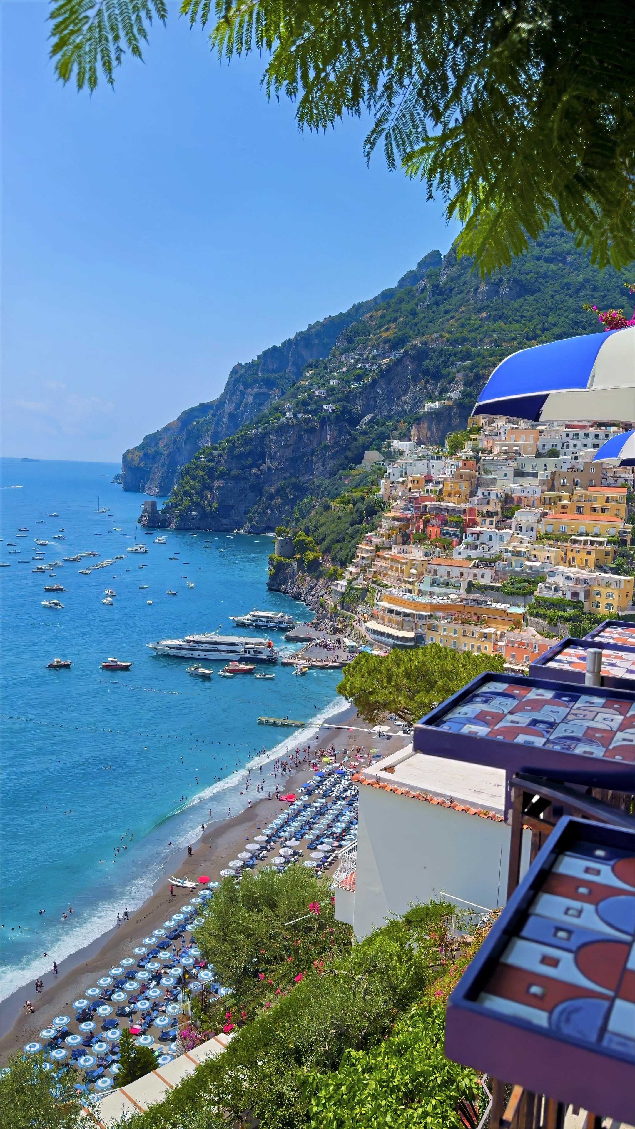 A curving pebble beach lined with umbrellas and boats, seen from above between pine branches.