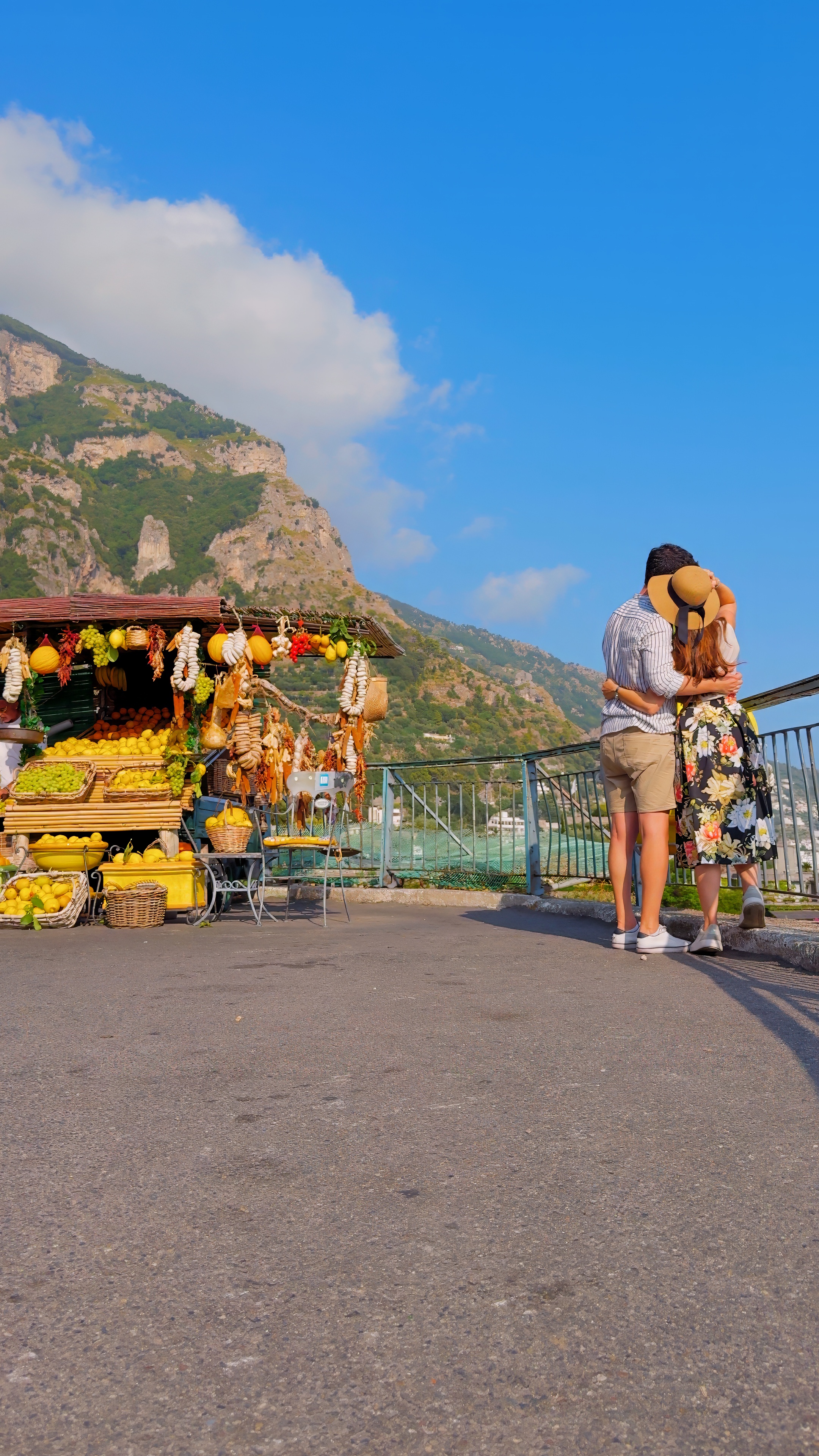 A couple kissing beside a flower stall on a coastal road, with cliffs behind