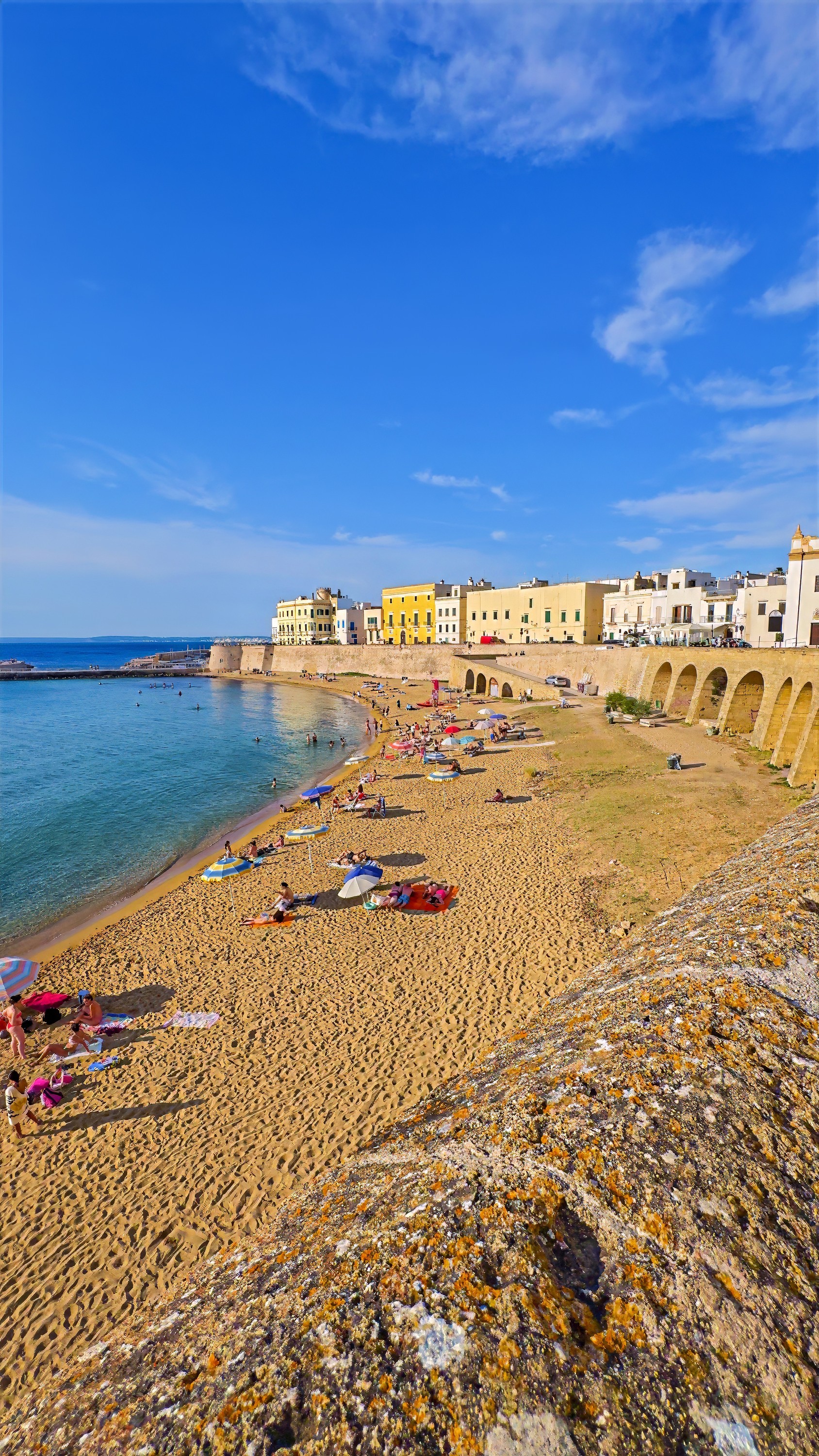 A small sandy beach below the old stone walls of a seaside town, with clear turquoise water.