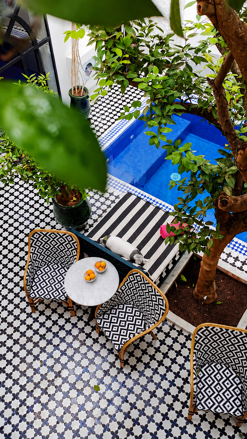 A table set on black-and-white tiles beside a blue pool, framed by leaves