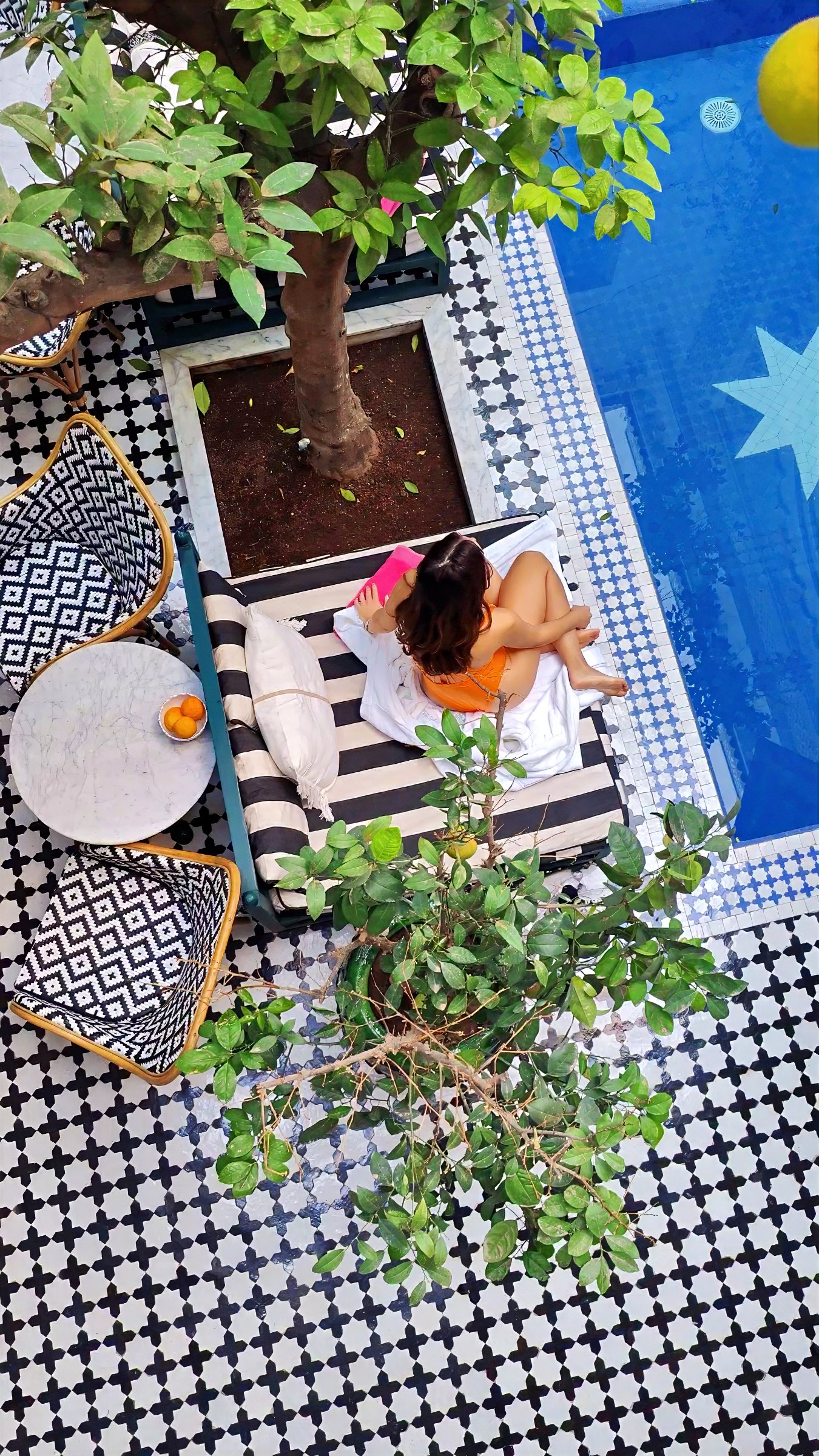 Seen from above, one of us resting on a daybed beside the courtyard pool, among potted plants