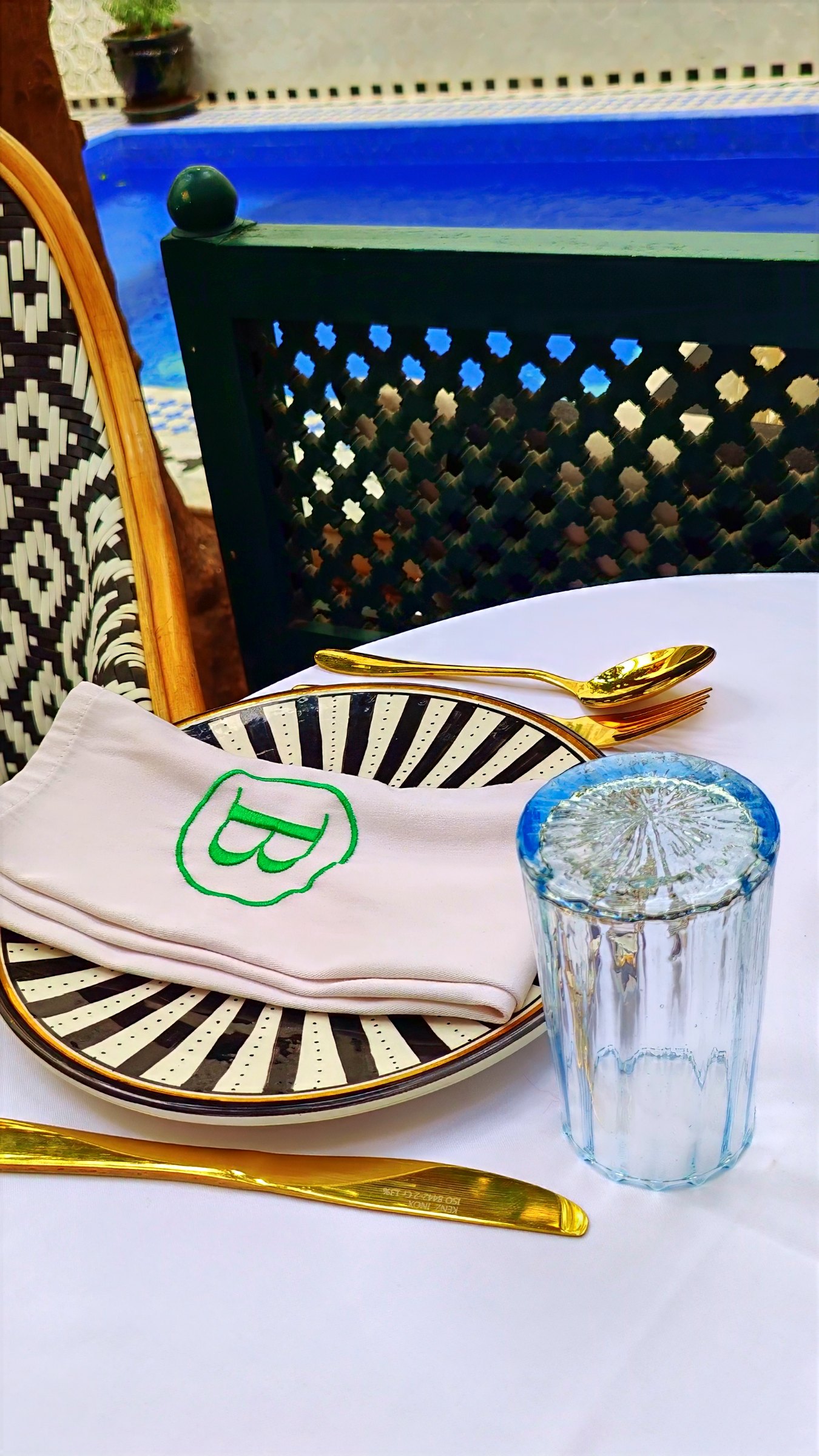 A lunch place setting beside the pool: a napkin embroidered with a green B on a black-and-white plate, a glass and gold cutlery