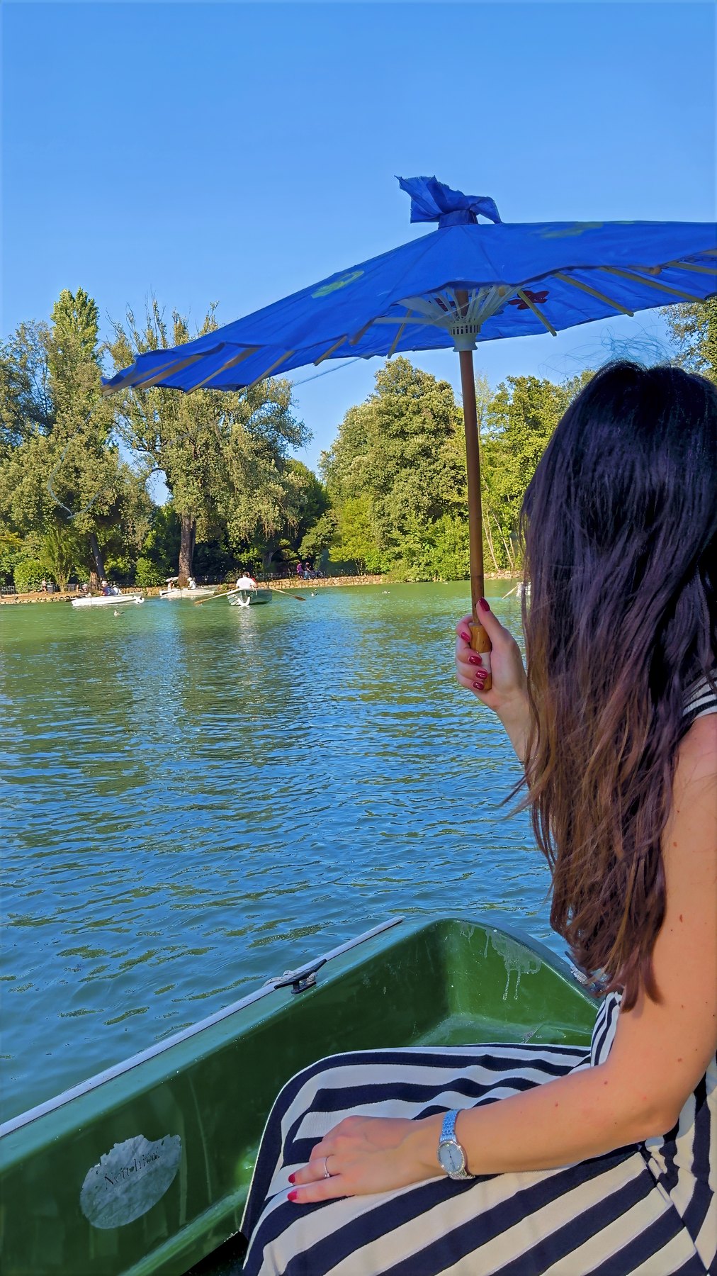 Barbara under a blue parasol in a green rowing boat on a small lake ringed by trees.