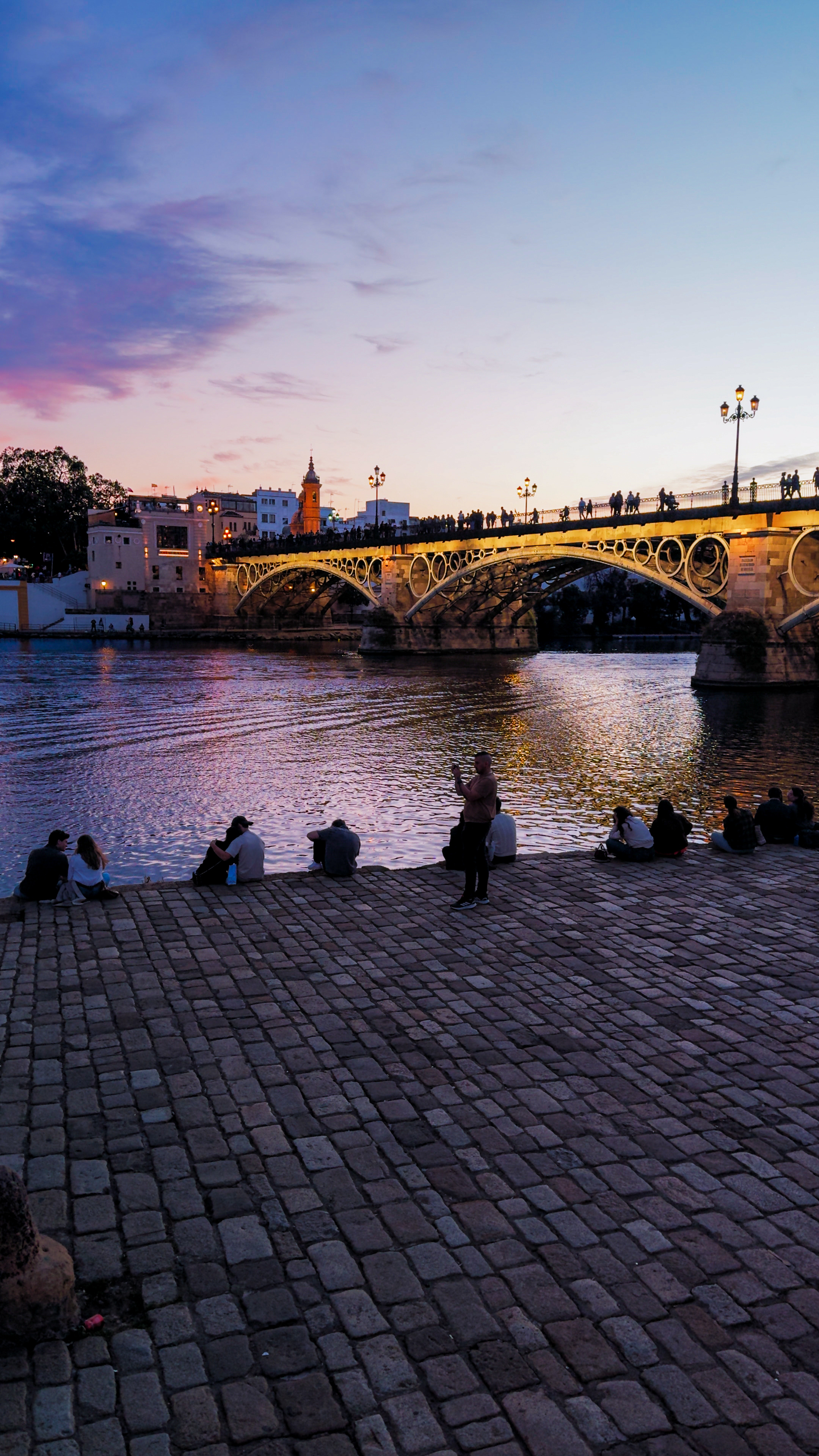 The Puente de Isabel II and the Triana riverfront at dusk, with people sitting on the city bank