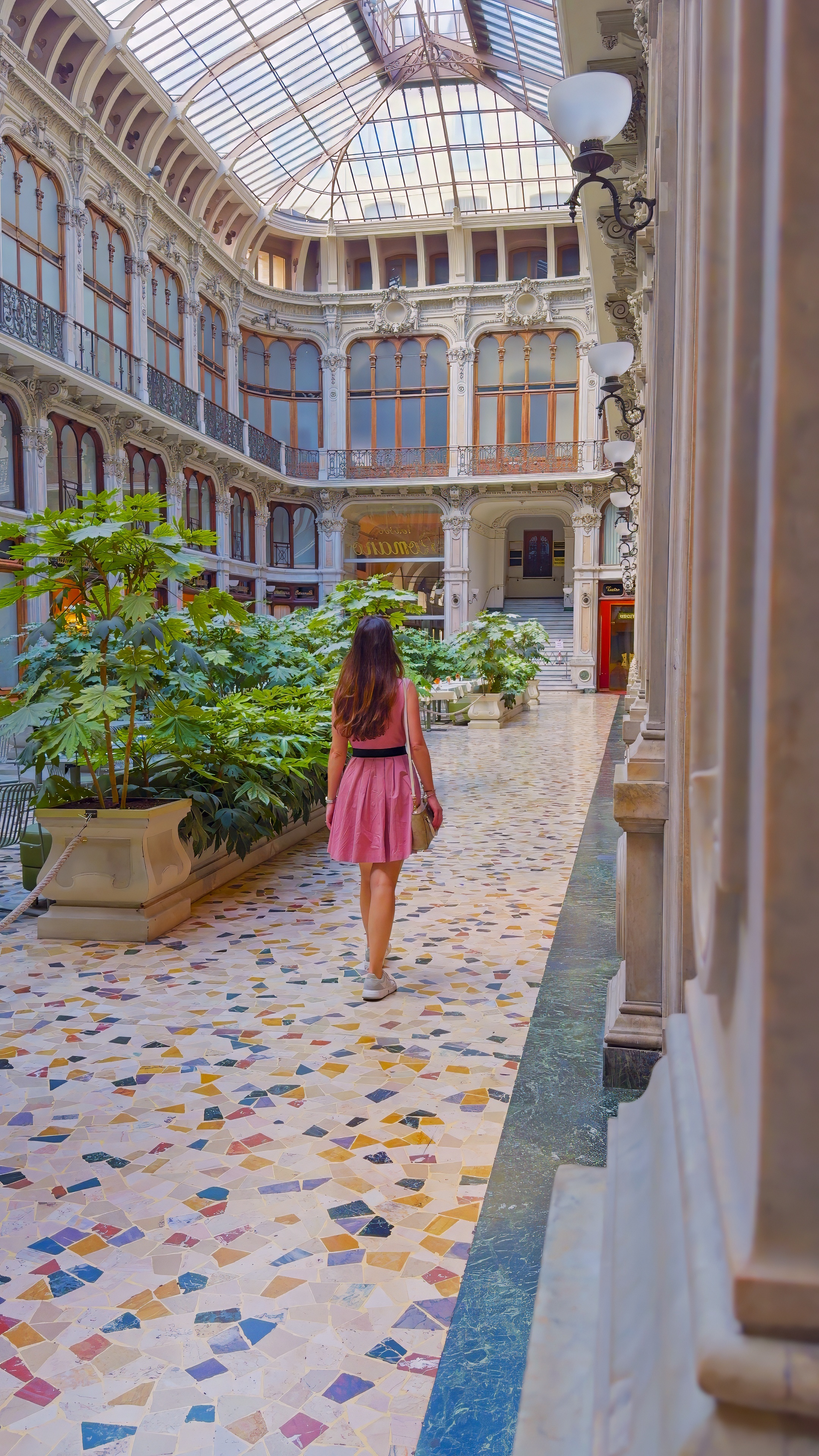 Barbara walking through a glass-roofed gallery with a mosaic floor and potted plants.