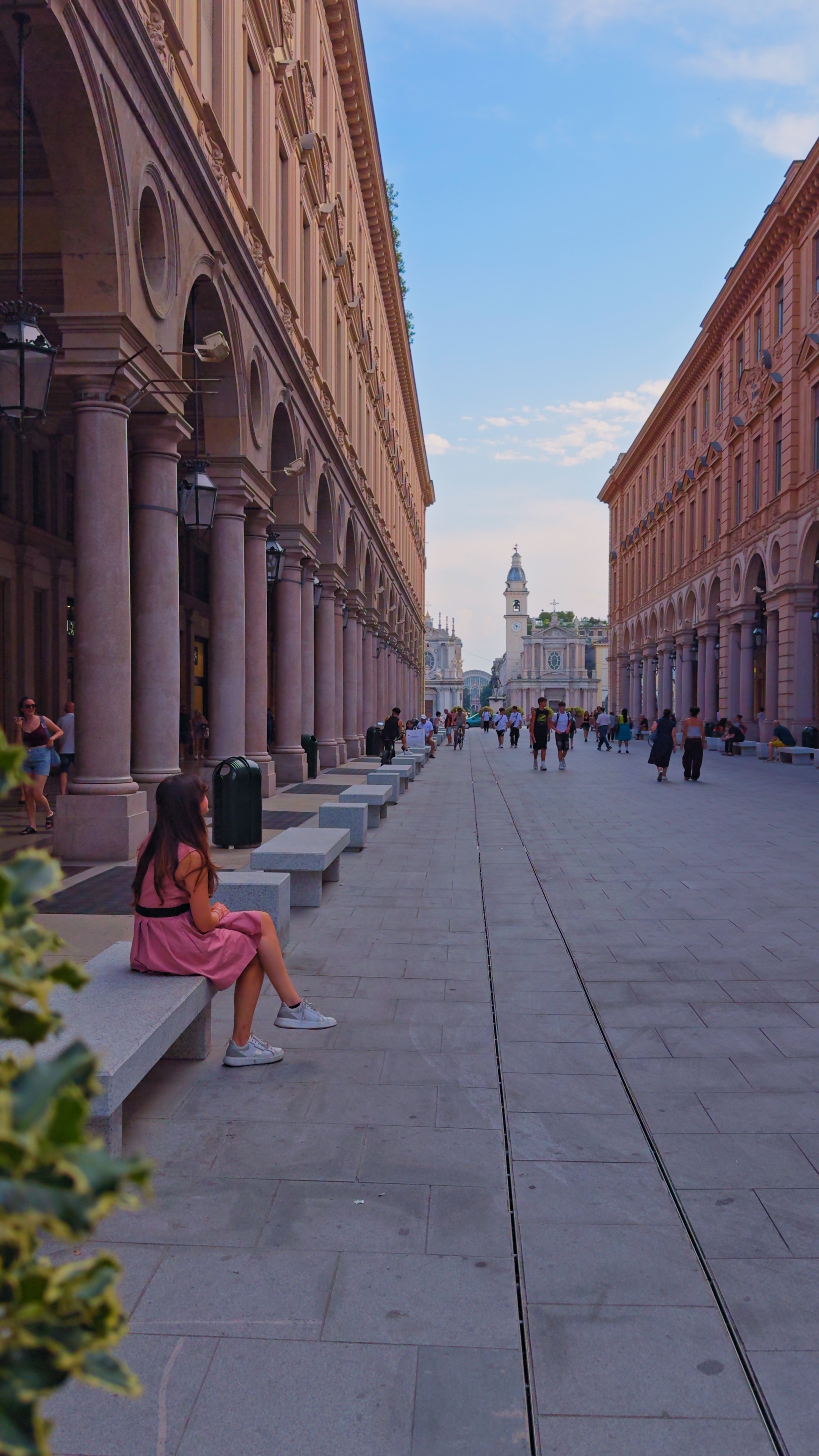 Barbara seated on a stone bench beneath a long arcade, a church at the end of the street.