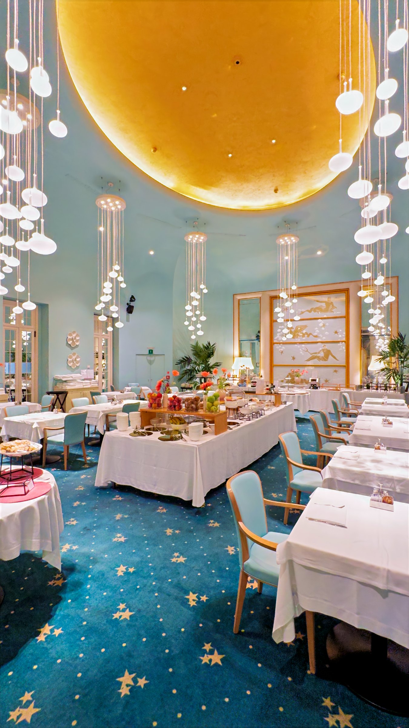 The breakfast room, with a gilded dome in the ceiling, glass chandeliers, blue carpet and tables laid in white