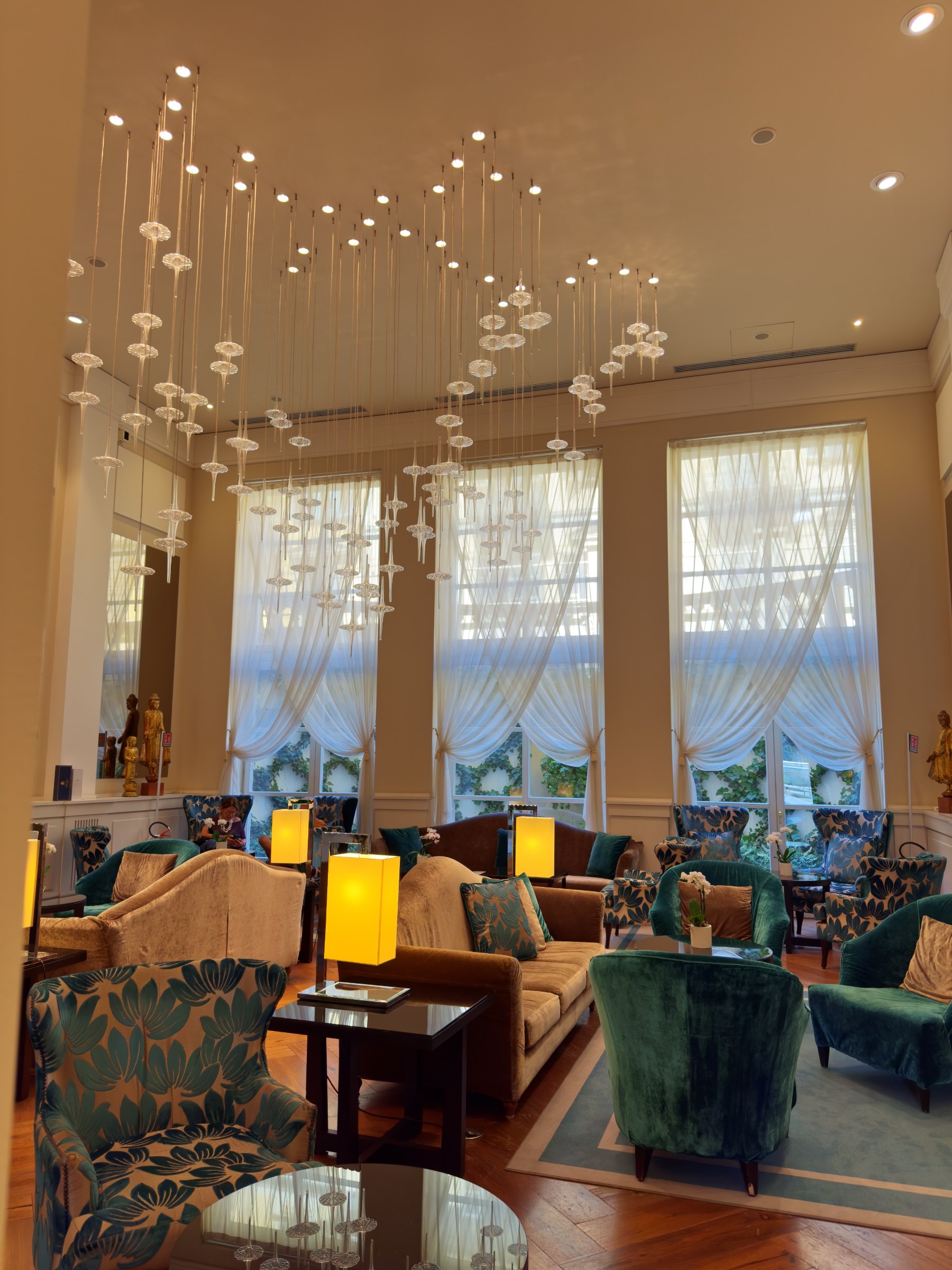 The ground-floor lounge, with teal velvet armchairs, brown sofas, tall windows and hanging glass lights