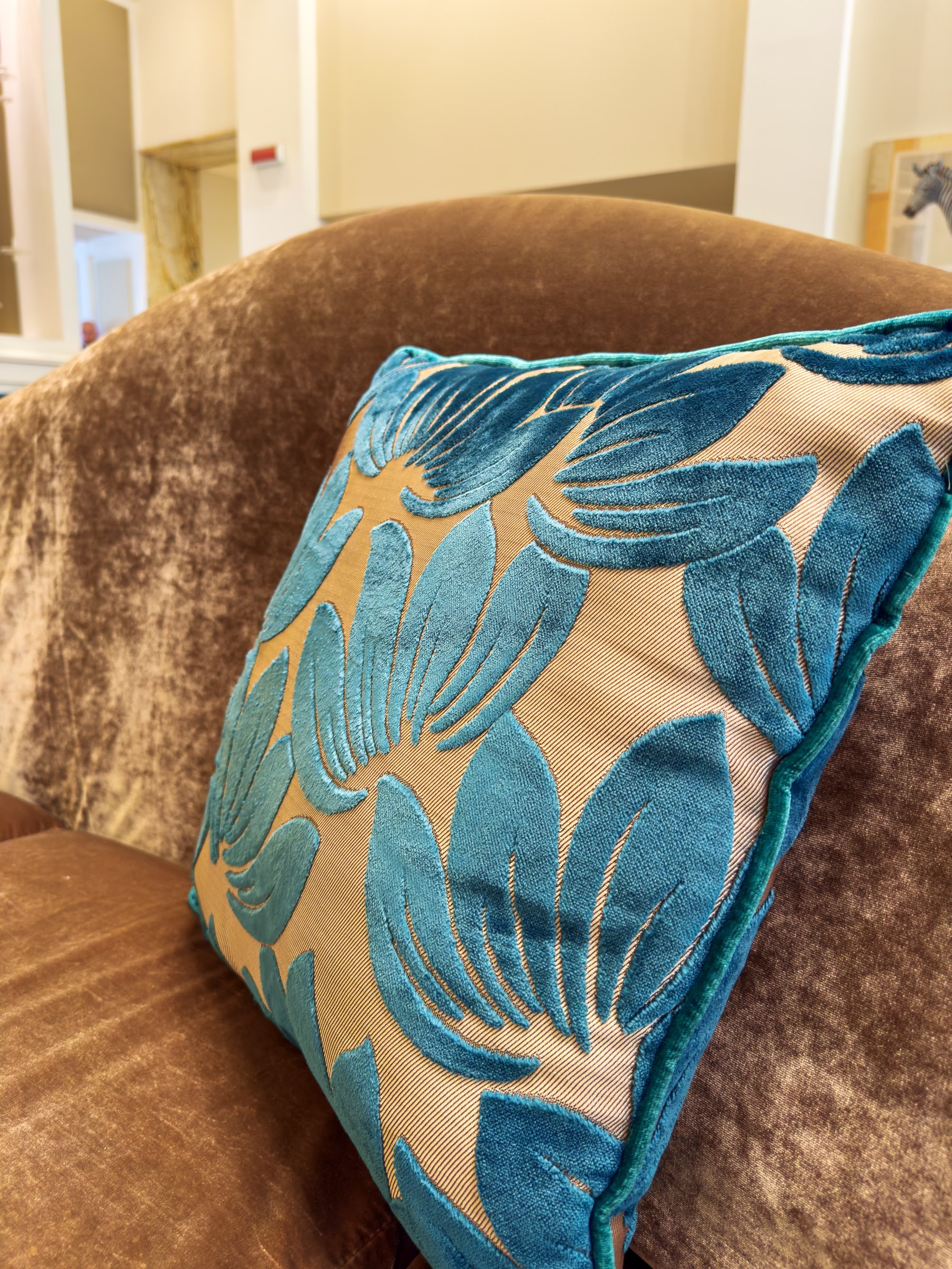 A teal velvet cushion printed with gold animals on a brown velvet sofa