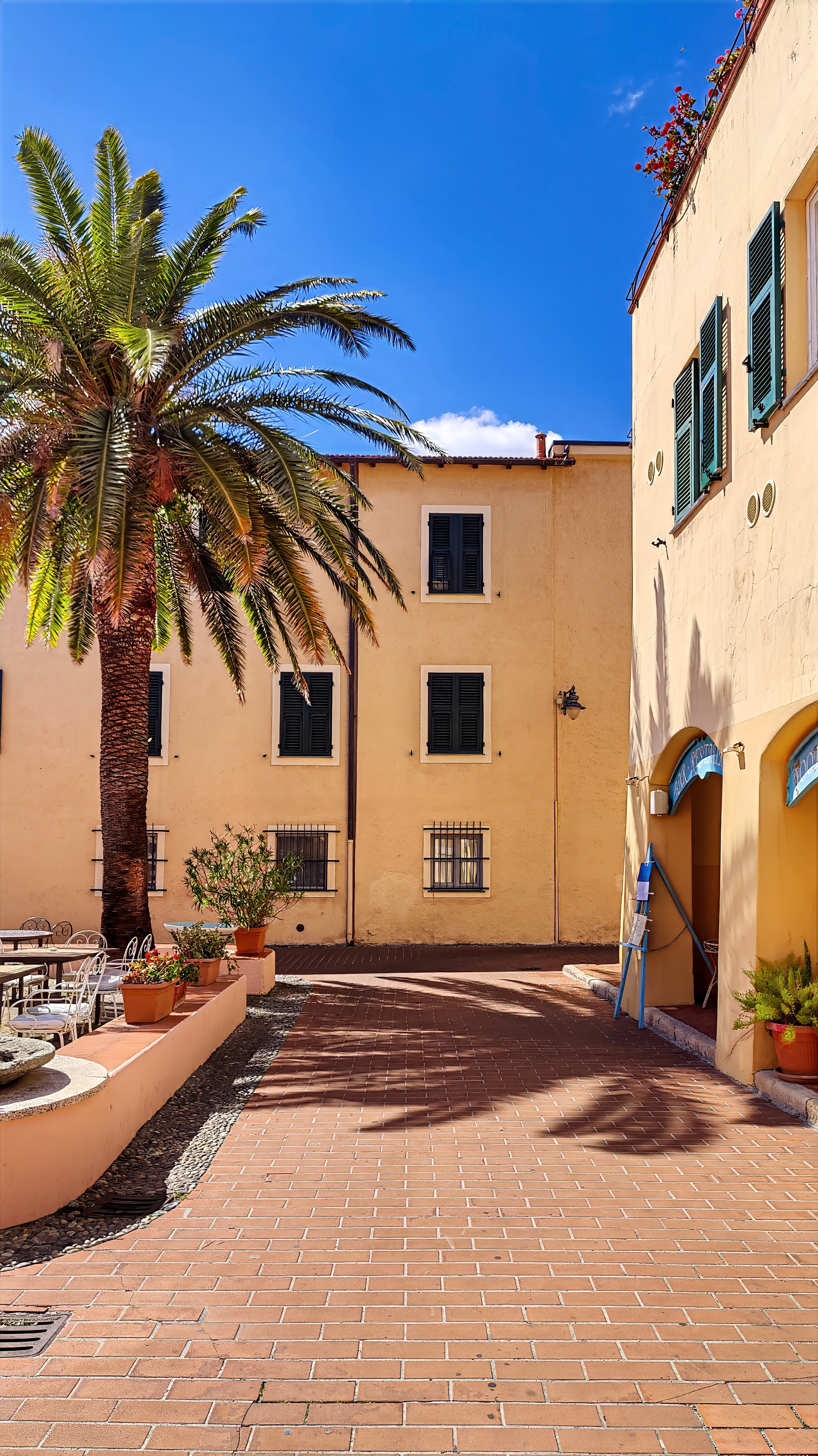 A small square in the borgo with a palm tree, ochre façades with green shutters and a brick pavement.