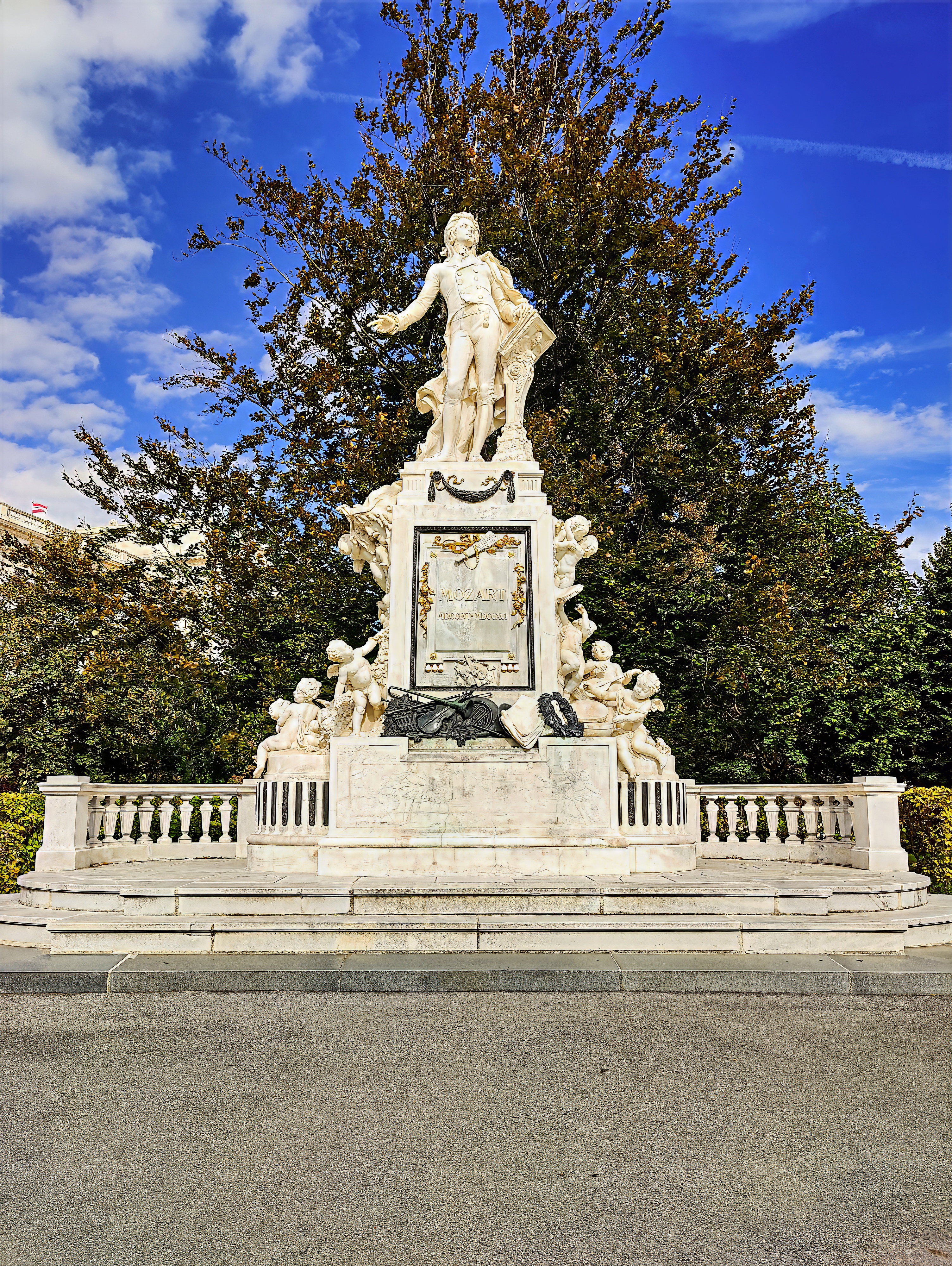 The white marble Mozart monument in the Burggarten, on a balustraded plinth in front of tall trees.