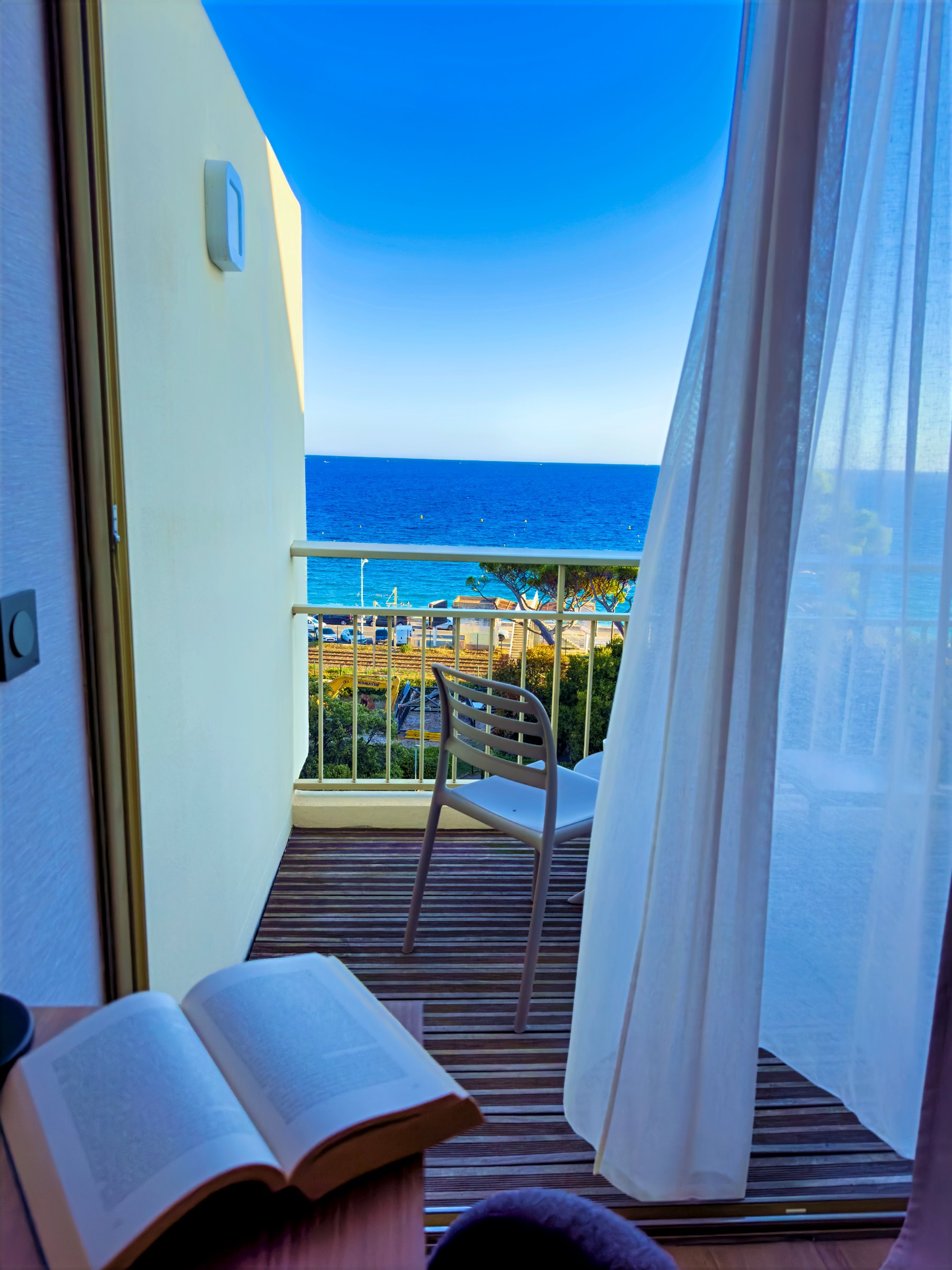 An open book on a table by a balcony door, with sheer curtains and the sea beyond