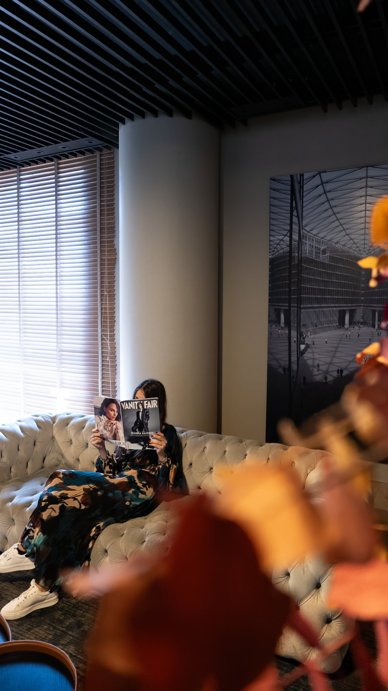 A woman reading a magazine on a tufted sofa in a hotel lounge, beside slatted blinds and a large architectural print