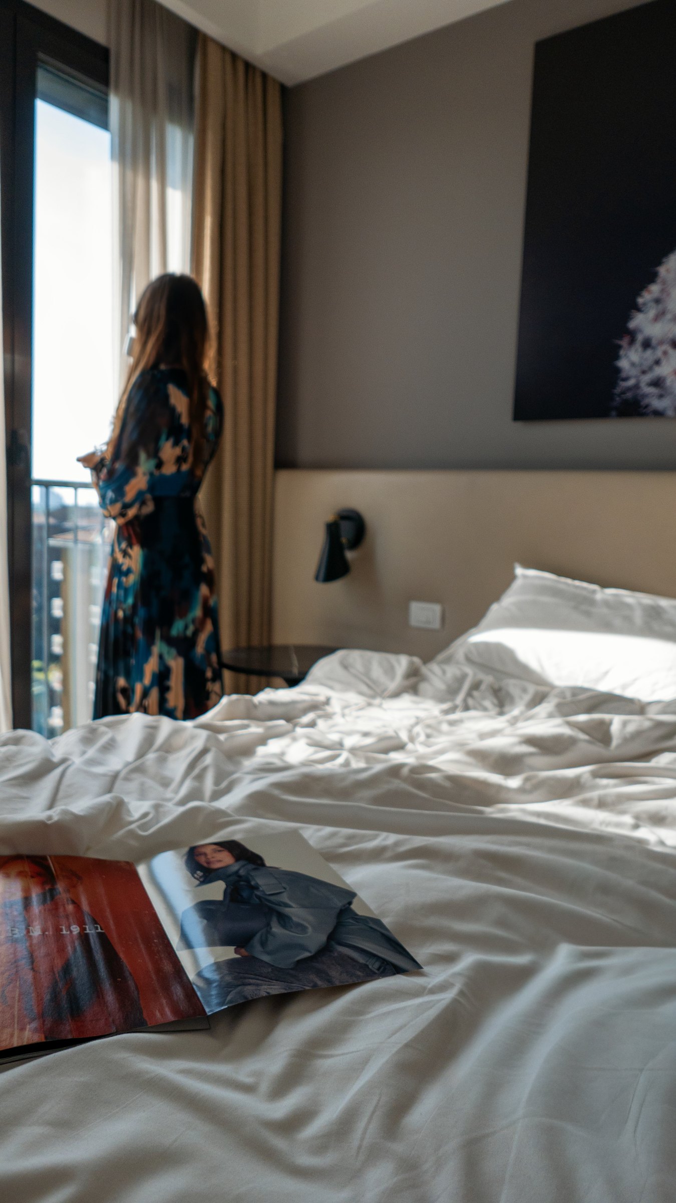 A hotel room in morning light: an unmade bed with an open magazine, and a woman standing at the window